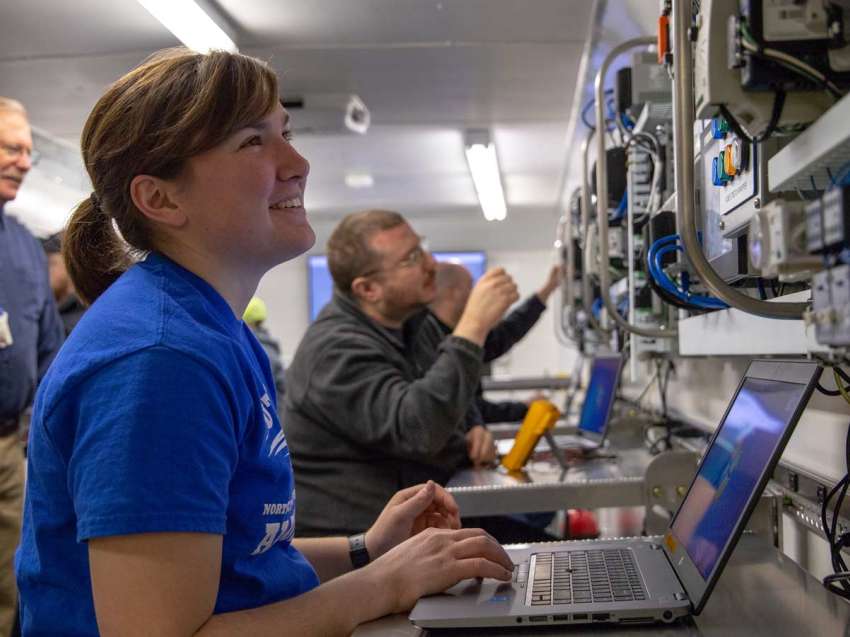 A student sits at a table in the Emerging Technologies Mobile Lab, as she works on a laptop and looks at controls mounted on the wall.