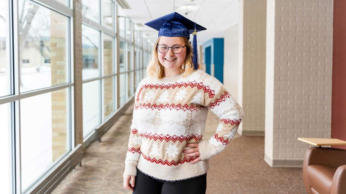 Molly Rydeski standing in an NTC hallway, wearing a festive holiday sweater and a graduation cap.