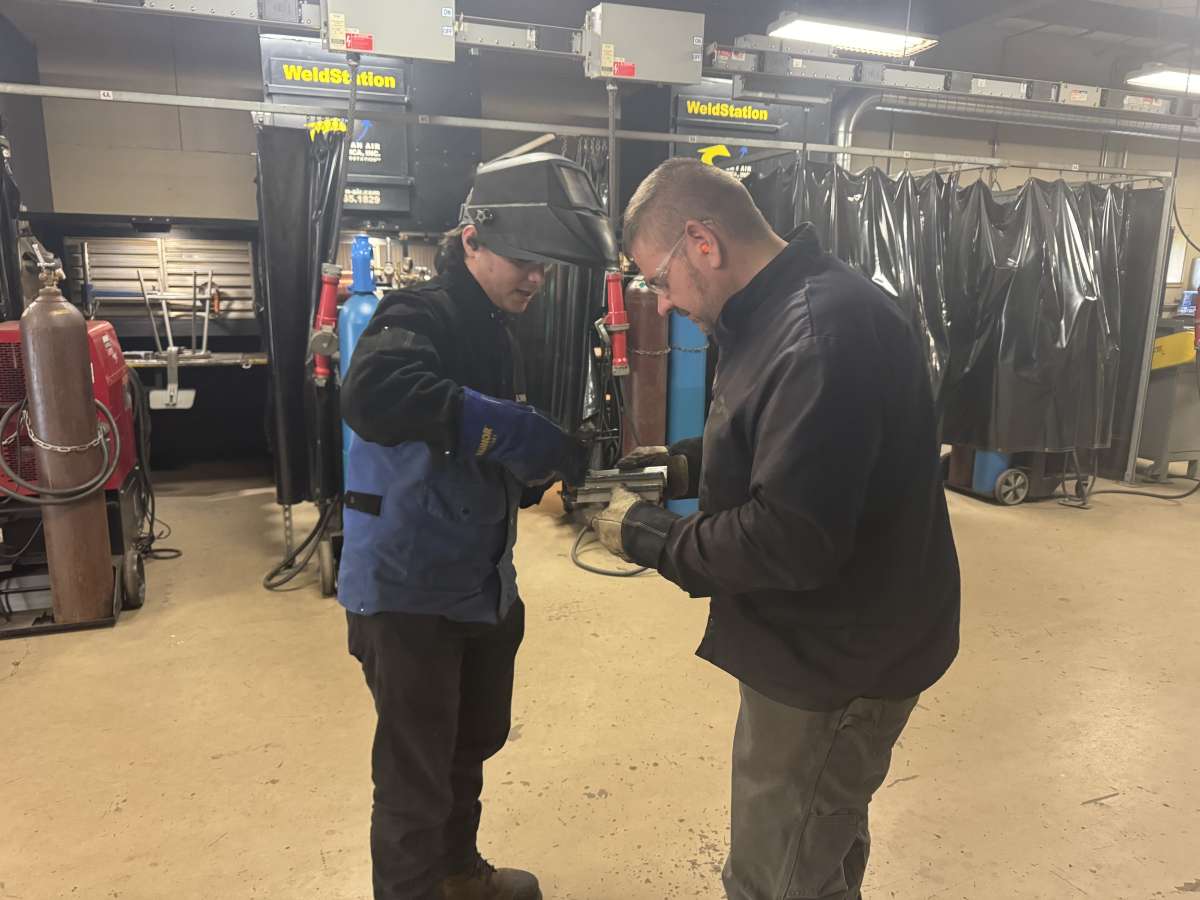 A welding instructor works one-on-one with a student surrounded by welding booths and industry-standard equipment.