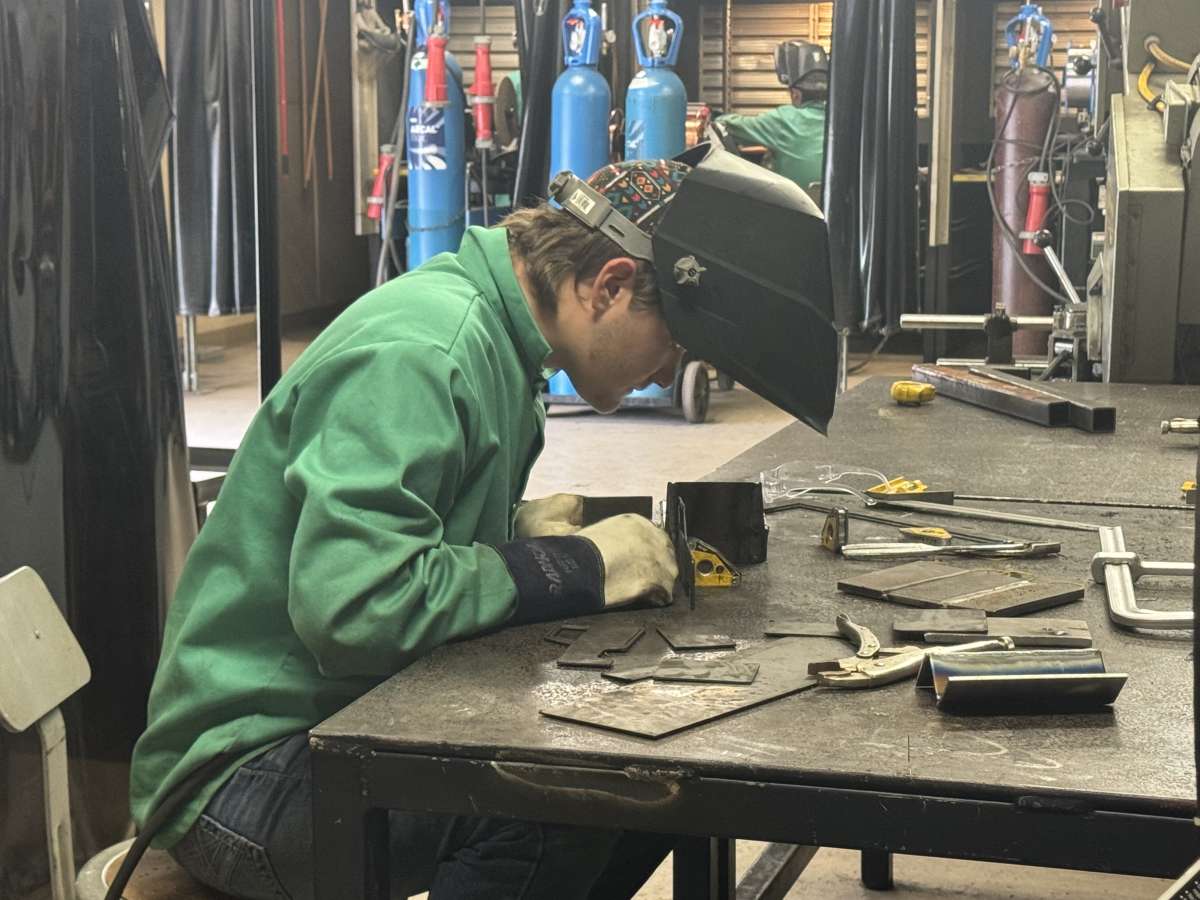 A welding student prepares metal components for fabrication at a workstation while wearing protective gear and a raised welding helmet.