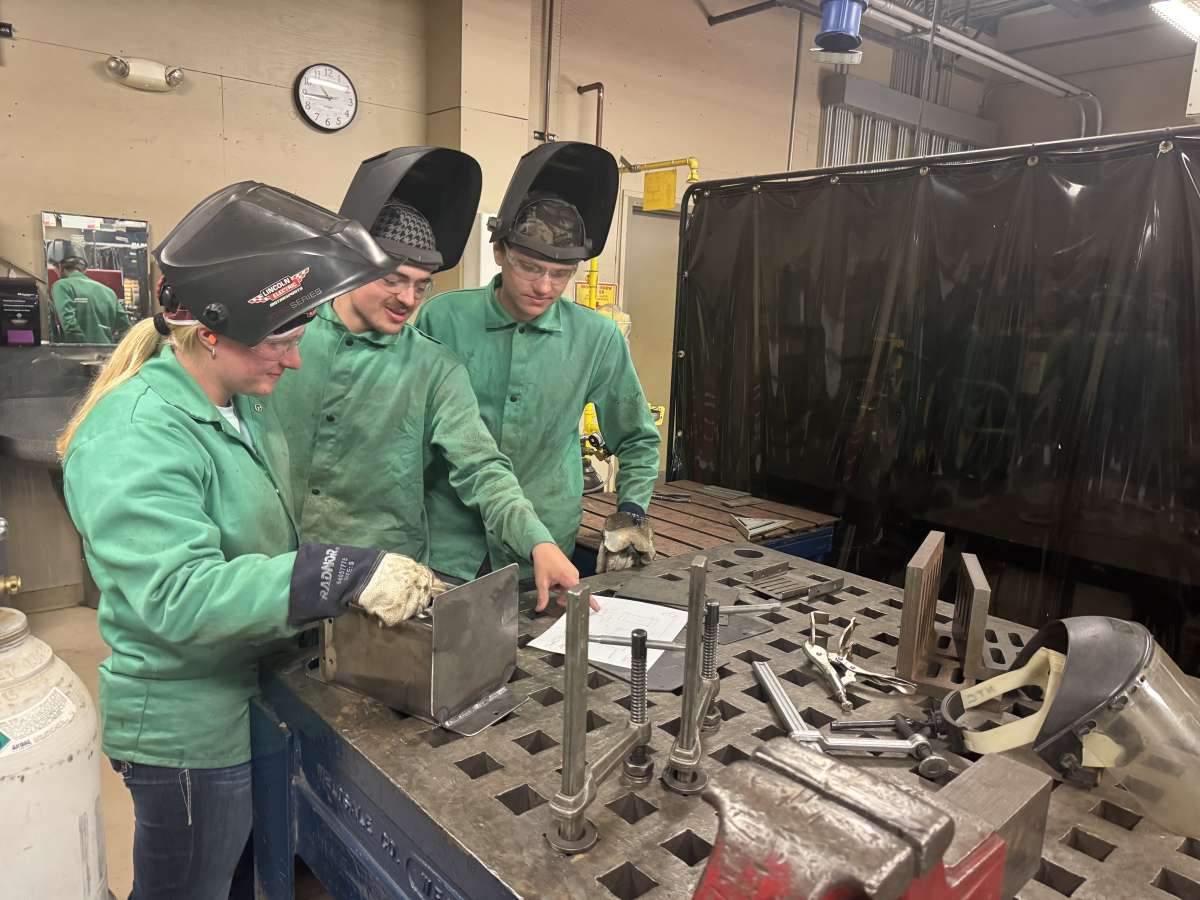 Three welding students gather around a fabrication table to review project plans before beginning work, wearing welding jackets, safety glasses, gloves, and raised welding helmets.