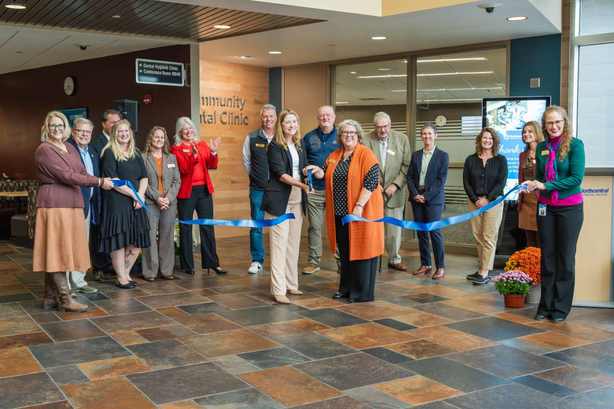 Group of people in a lobby cut a long blue ribbon with oversized scissors for a ribbon‑cutting ceremony.