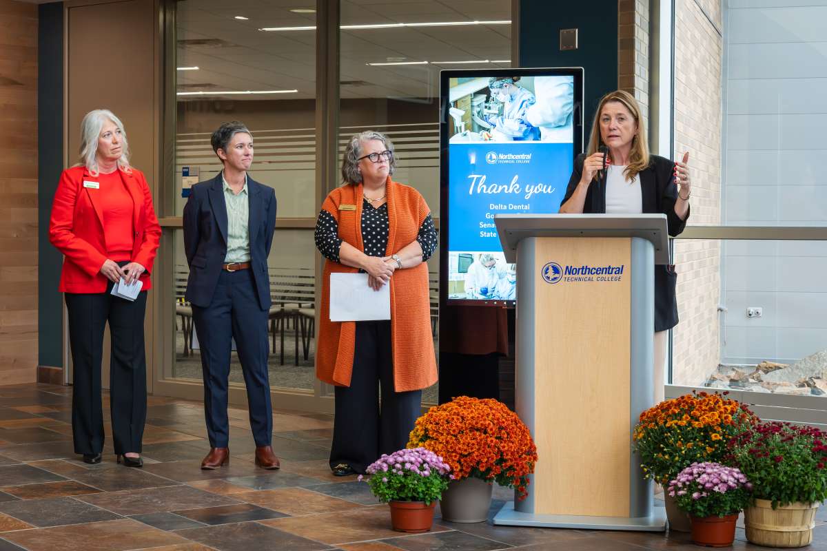 Four professionally dressed women in an atrium during an autumn ceremony; one speaks at a podium.