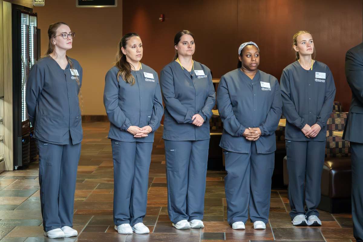 Five healthcare workers in navy scrubs standing side-by-side in a wood-paneled room with attentive expressions.