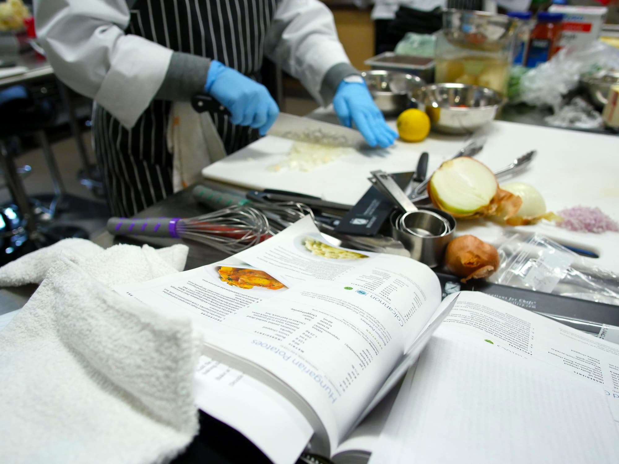 Close-up view of a busy kitchen work space. A student dices an onion as other tools and ingredients are kept nearby.