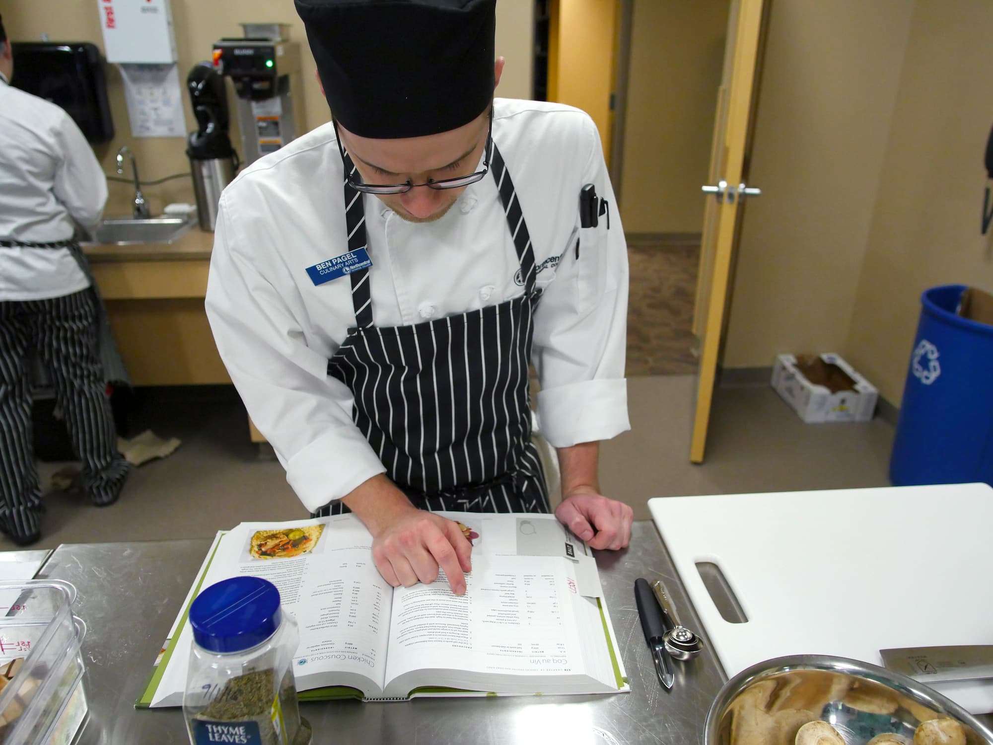 A student at a stainless steel kitchen work space scans a cookbook.