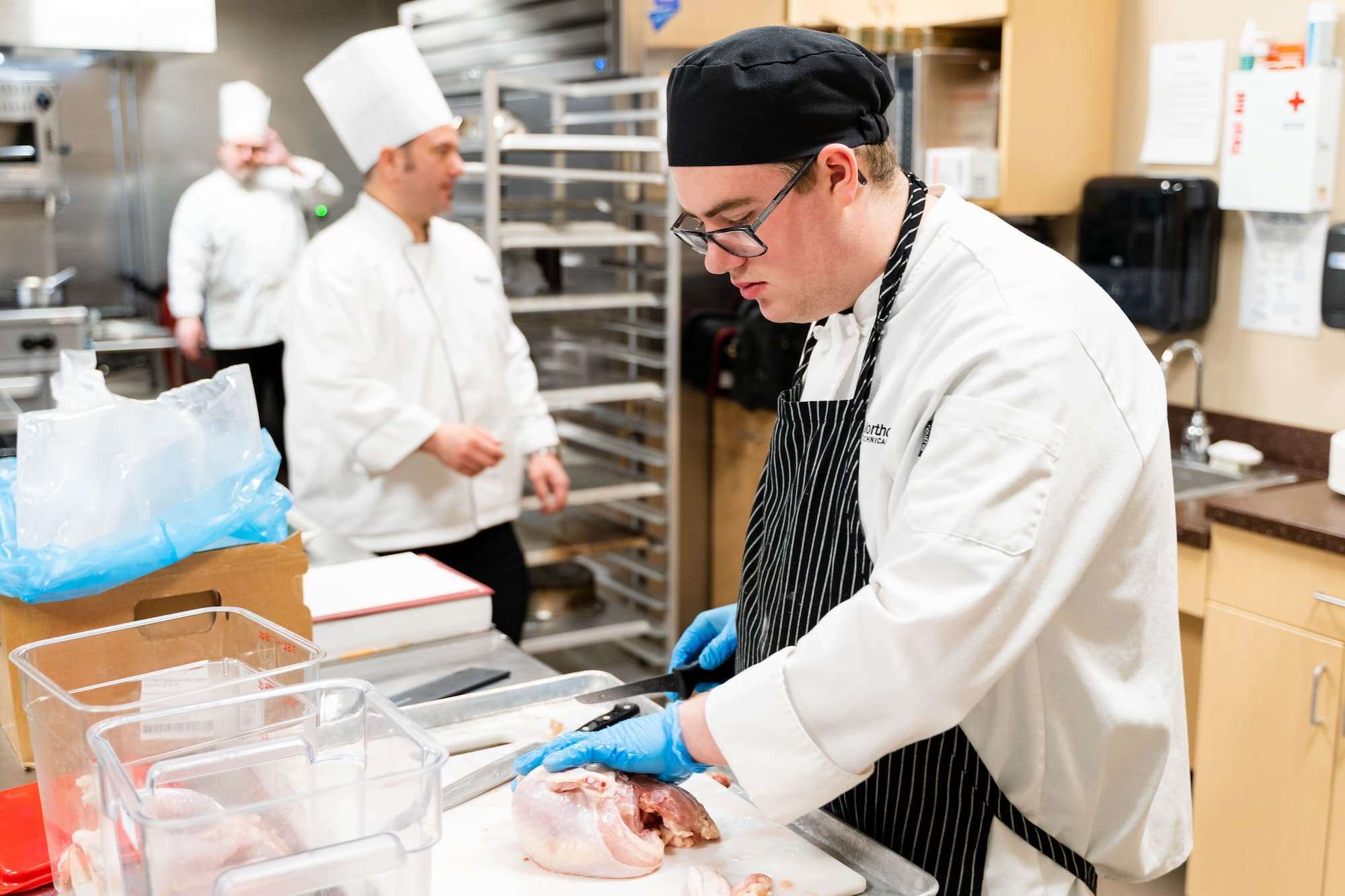 A student switches to a small carving knife to finish dividing a raw, whole chicken.