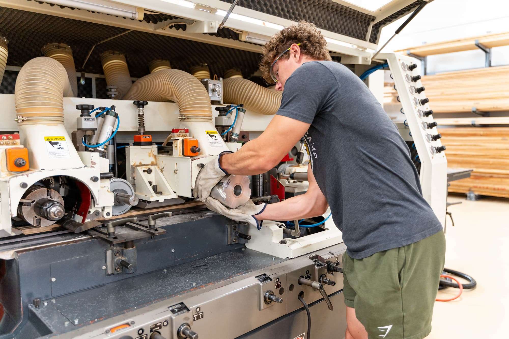 A student wearing thick gloves and safety glasses runs lumber through a moulder.