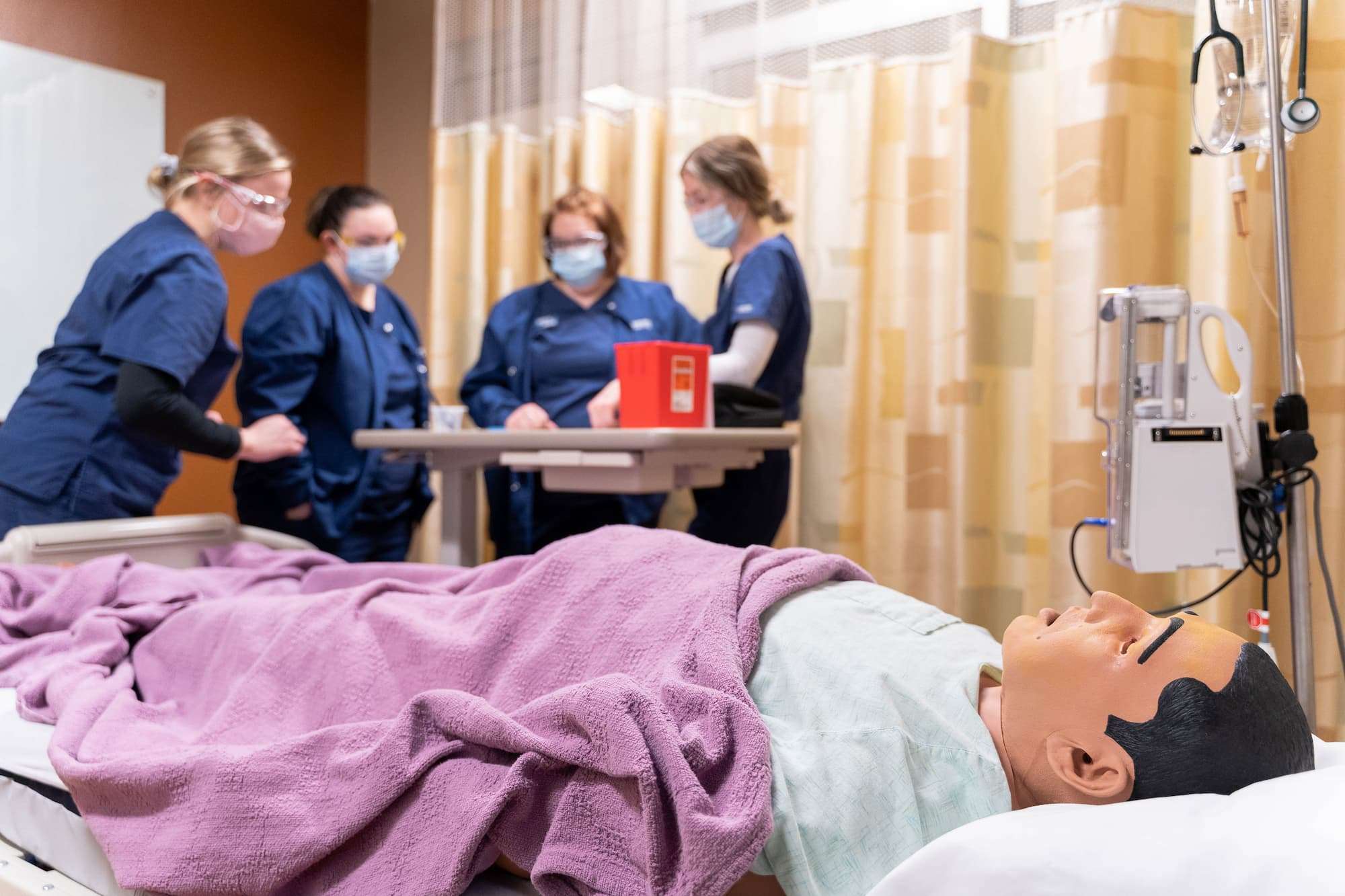 A close-up shows a mannequin in stasis as nursing students across the room discuss treatment.