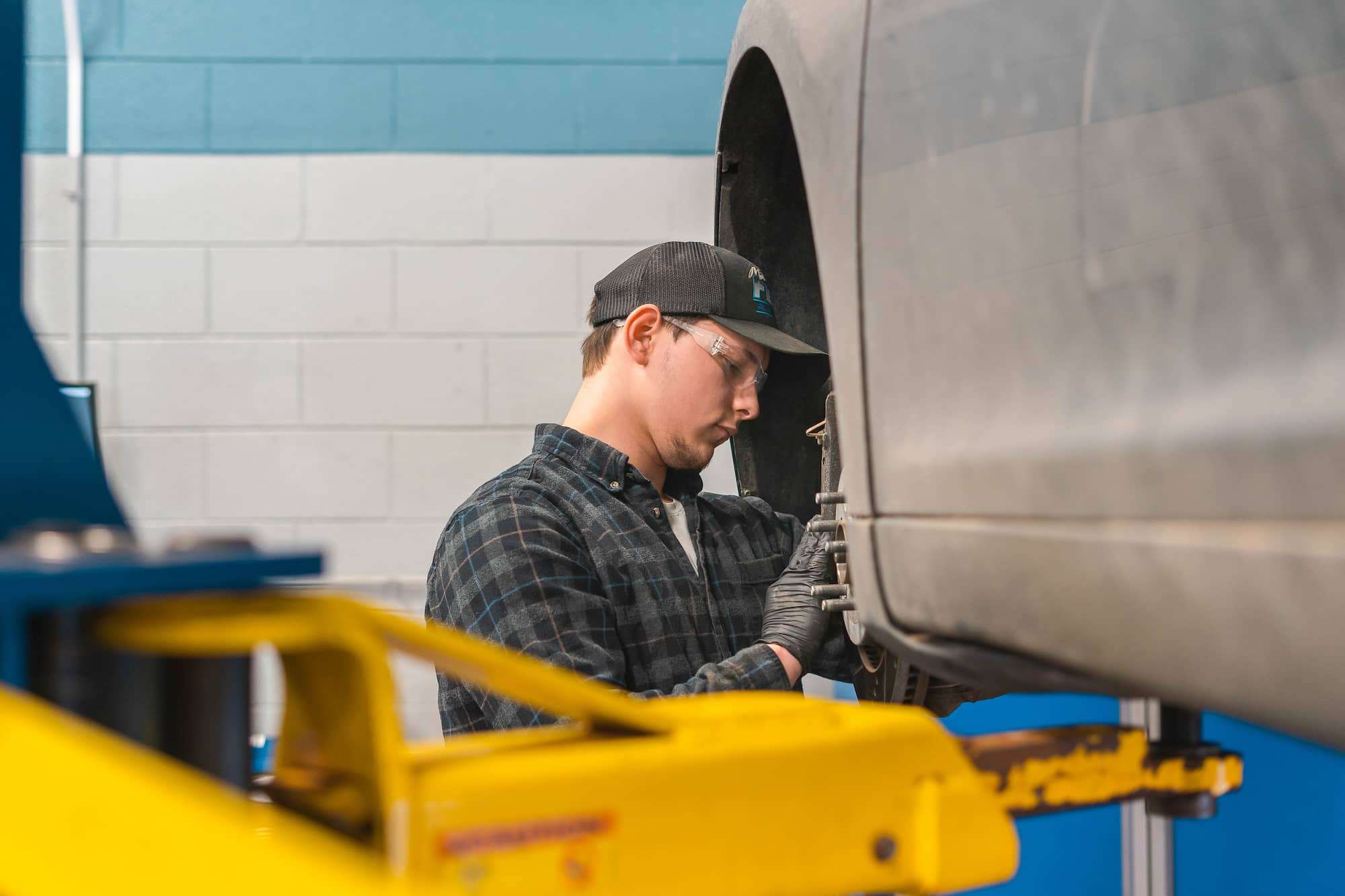 A man stands near the wheel well of a car that's been elevated on a hydraulic lift to eye level. The tire has been removed so that the brake pads and caliper can be inspected and repaired.