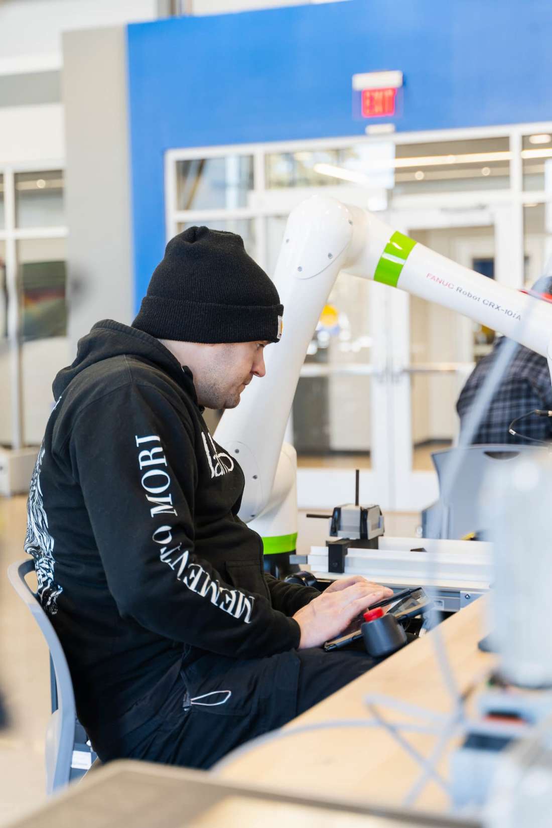 Profile view of a student wearing a black hat and hoodie, watching the operations of an industrial robot.