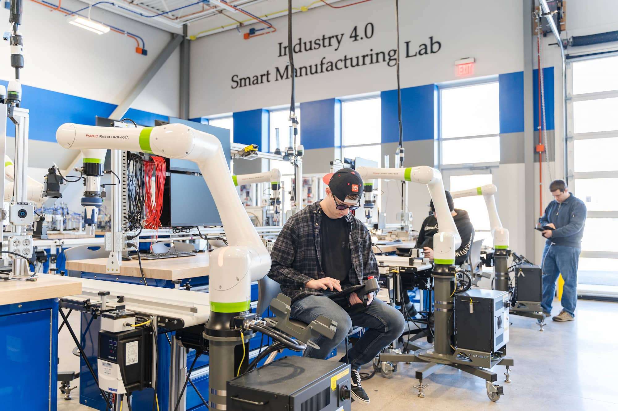 A wide-view shows NTC's Smart Manufacturing Lab, where three students are each configuring their own manufacturing robot.