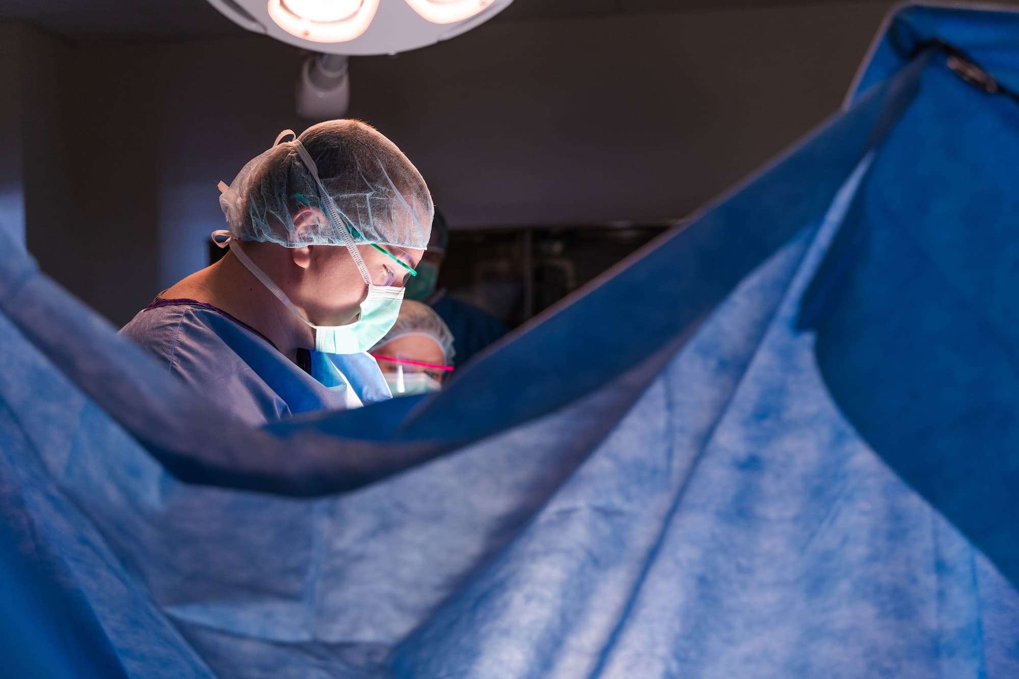 Lit by only a surgical lamp, a student wearing a medical gown, surgical mask, hair net, and safety glasses conducts an operation. His hands are obscured by a surgical drape.