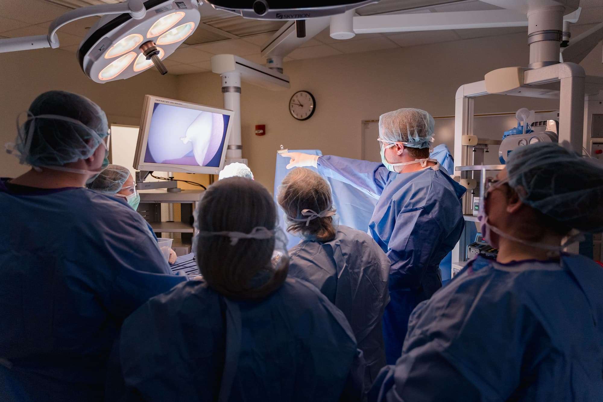 A student points out details from a surgical camera, displayed on an overhead monitor in the operating room.