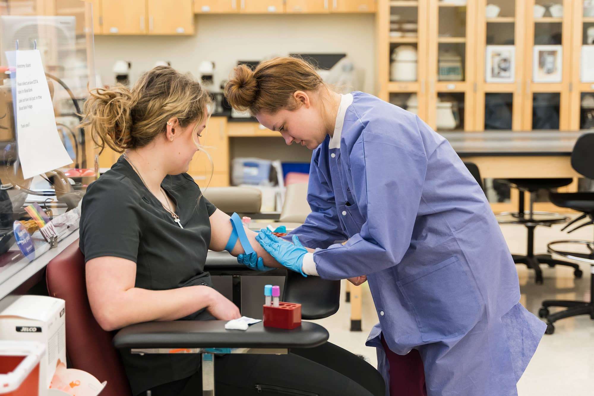 A view from the side of a Phlebotomy student placing their hand on a patient's forearm before drawing blood.