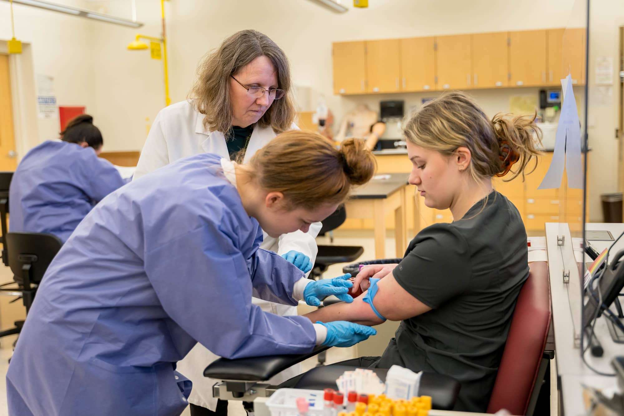 An instructor watches on as a student prepares to draw blood from a patient's arm.
