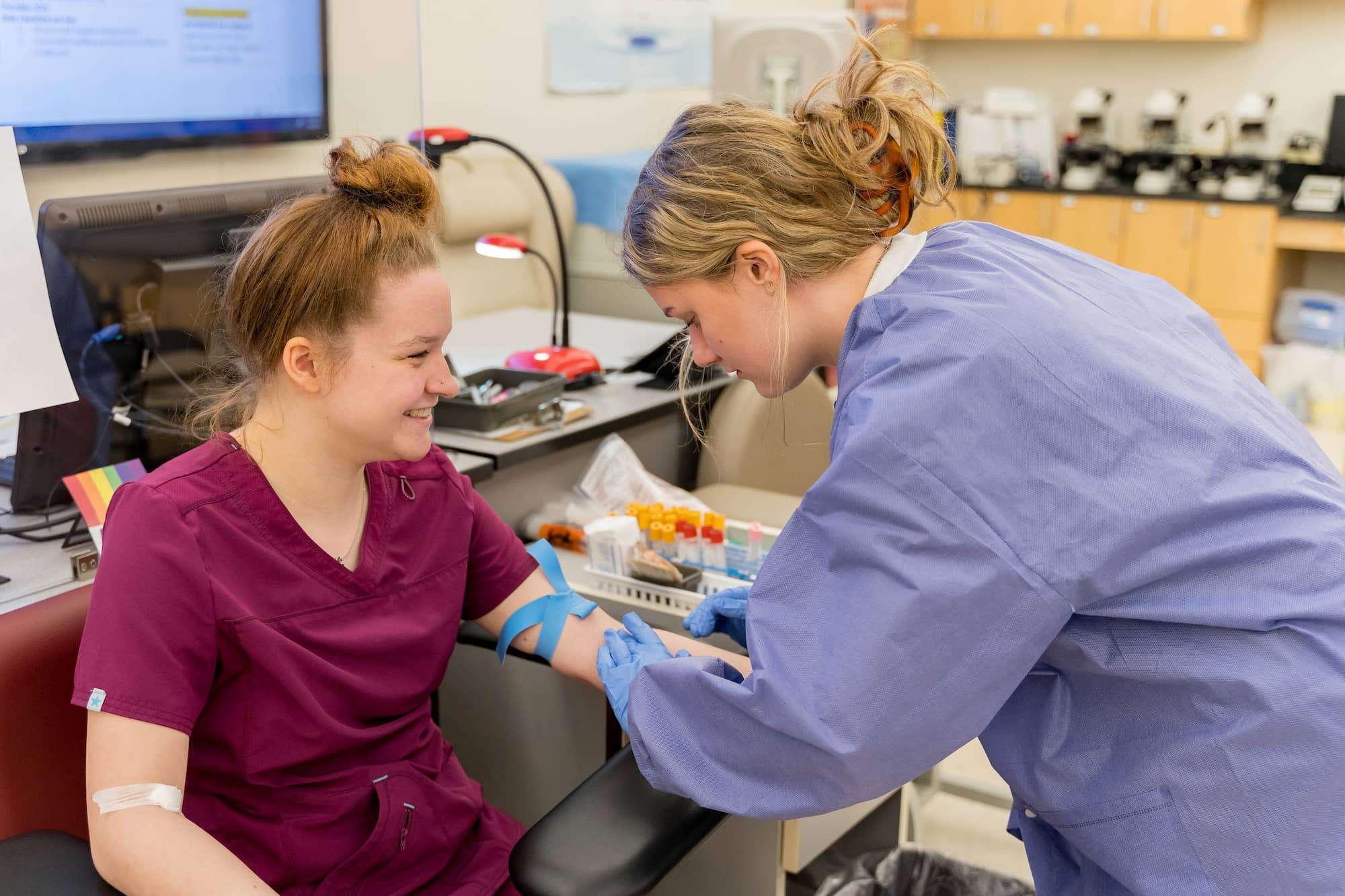 A student, acting as the patient, laughs as another student feels for the position of the patient's vein on her forearm.