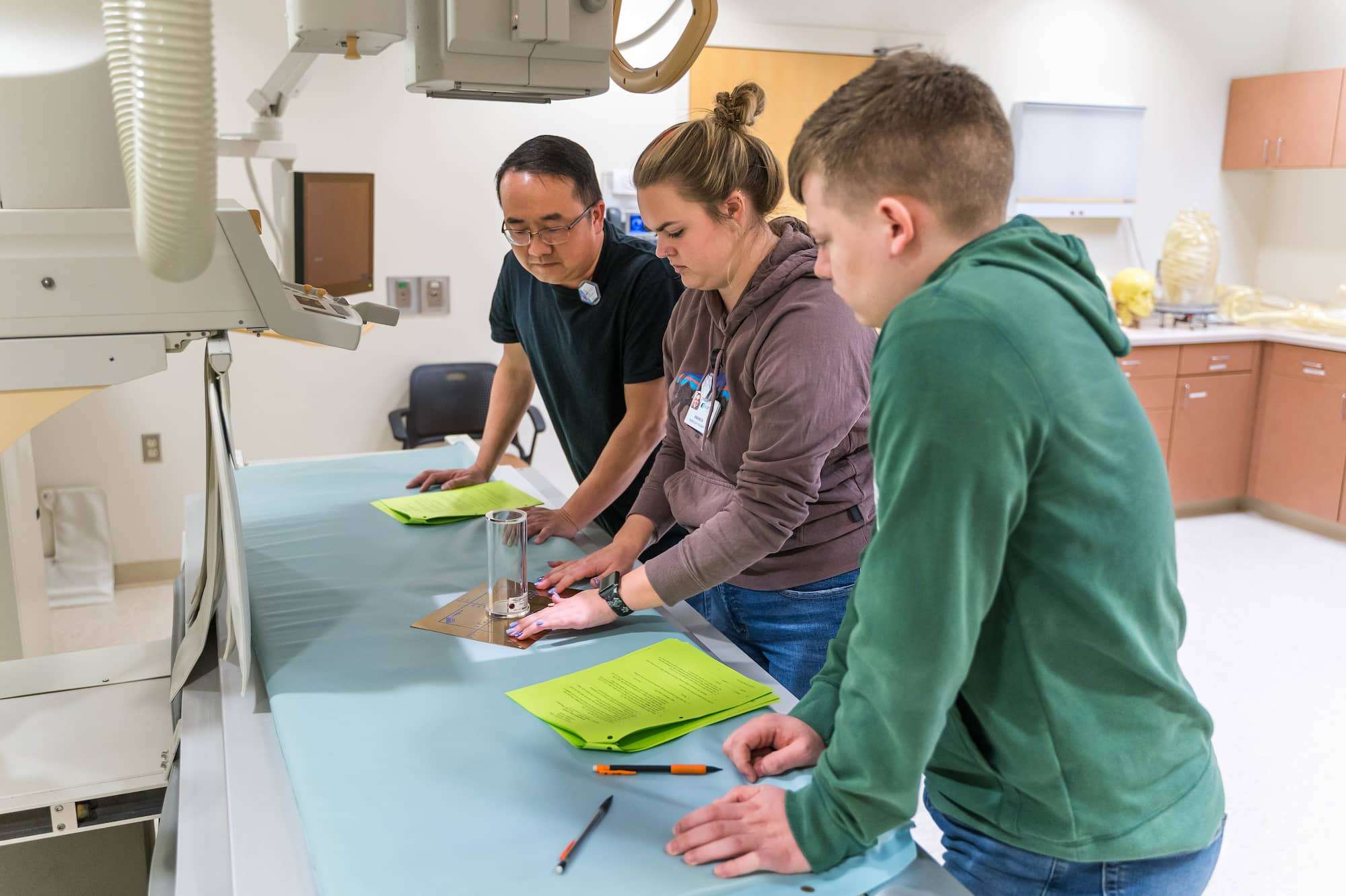 A group of students practice aligning a radiography system over a practice target placed on a patient bed.