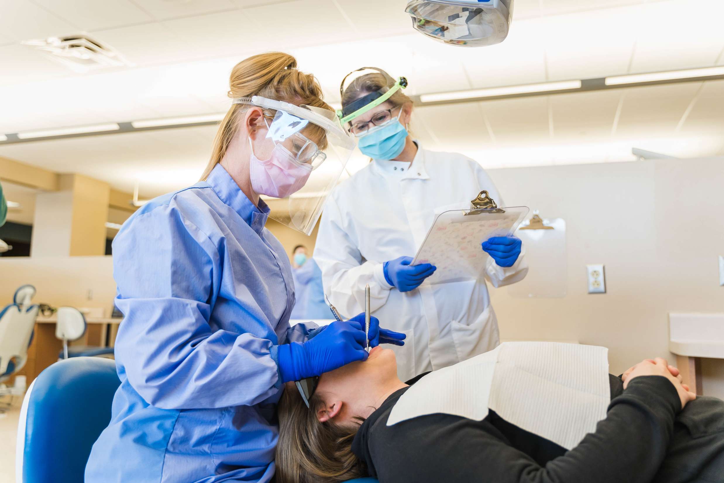 A dentist stands next to a learning student checking over her work with a clipboard.