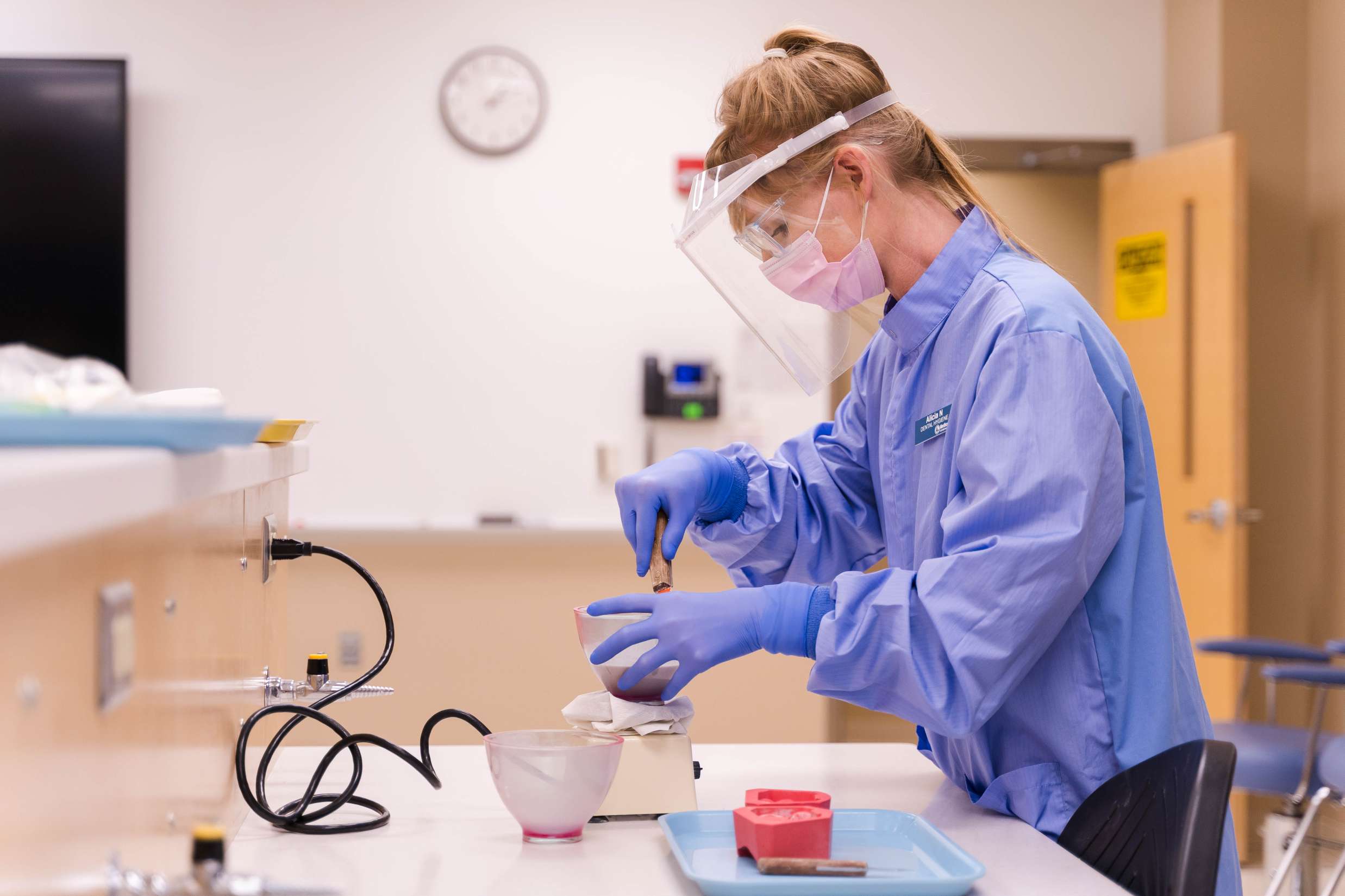 A dental hygienist wearing blue medical garbs and gloves working with dental molds inside of a lab.