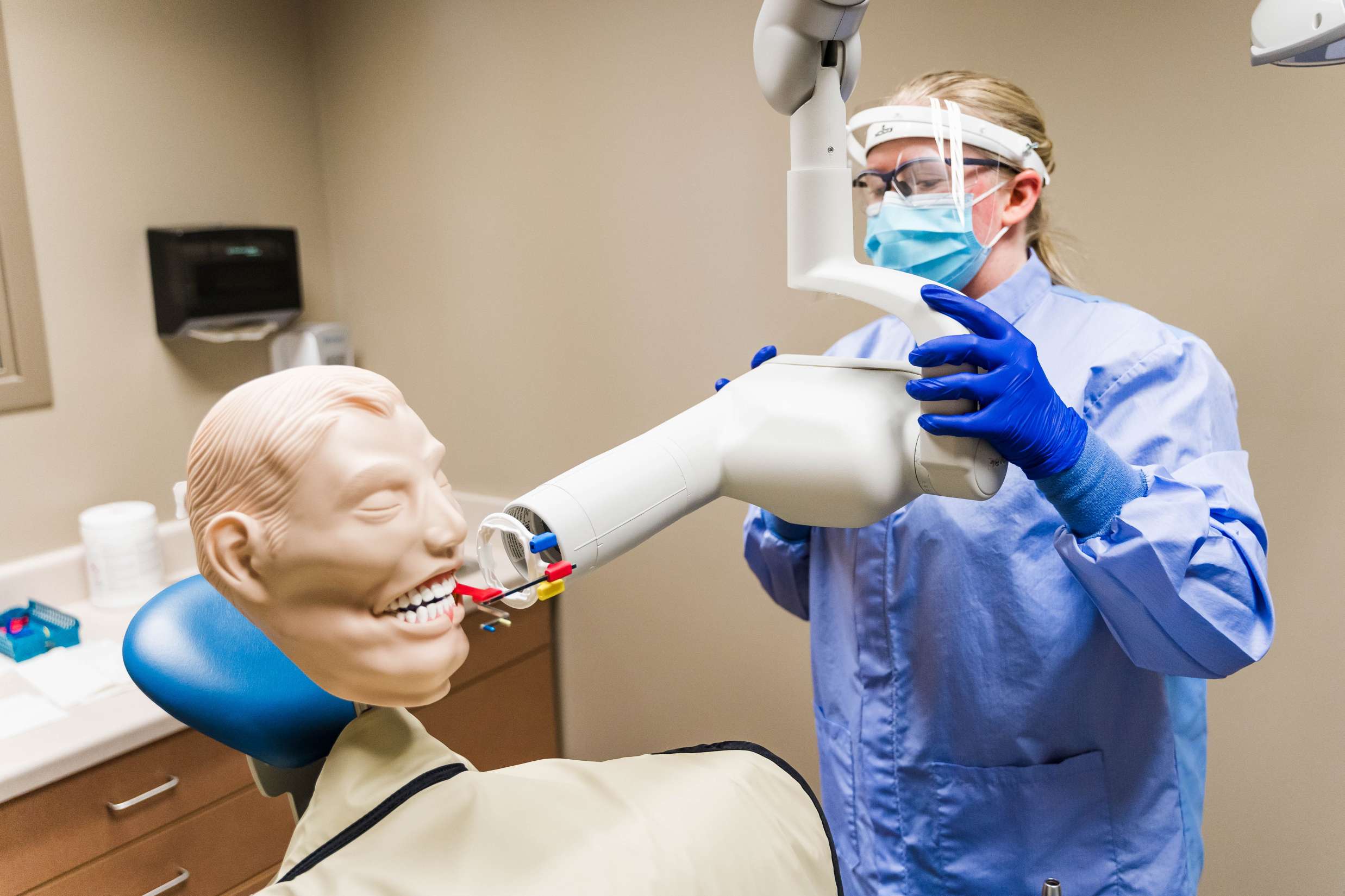 A dental hygienist wearing medical garbs and a face shield is using a mannequin to practice taking an x-ray of a mouth.