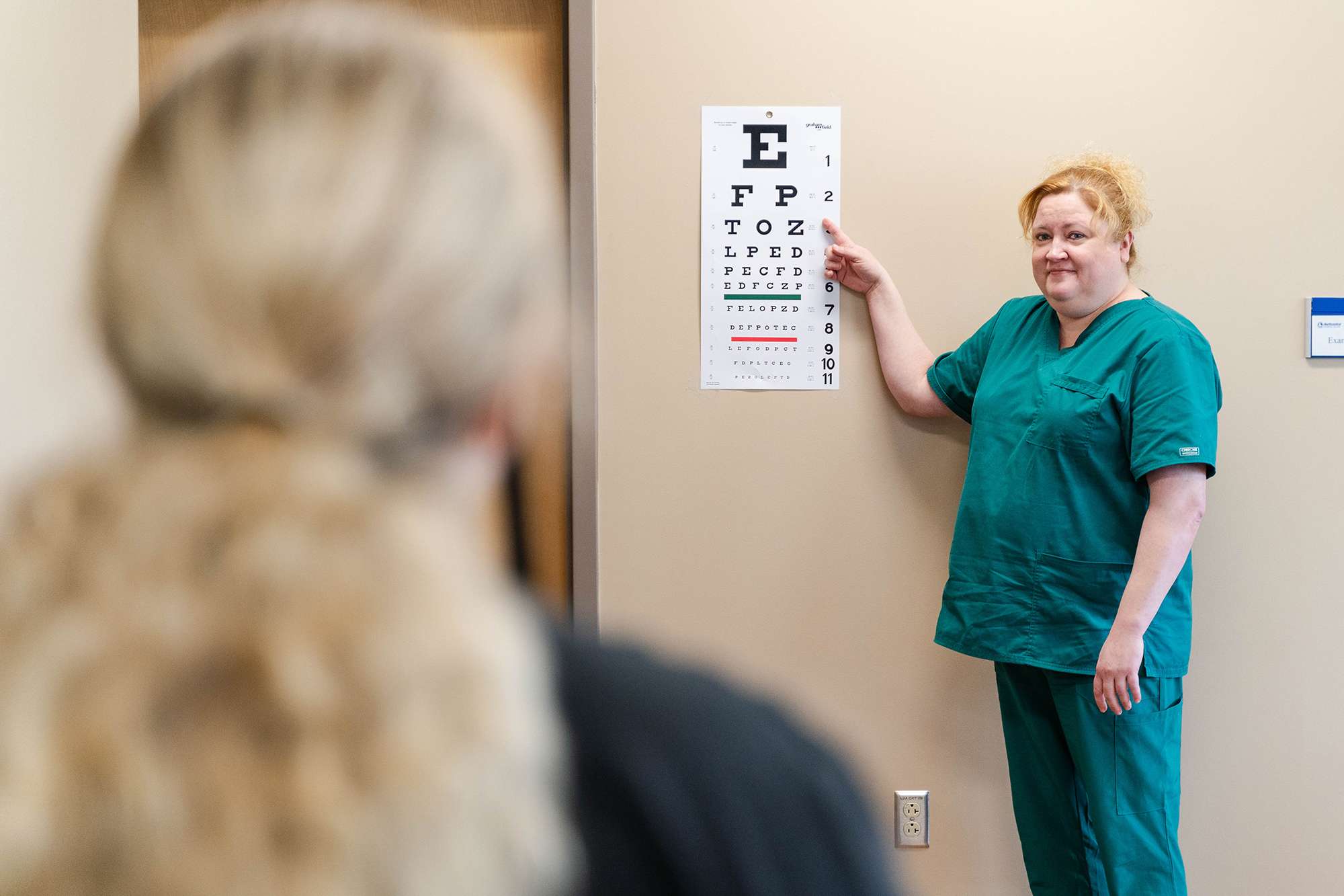 A Medical Assistant stands next to a Snellen eye chart as a patient reads the letters.