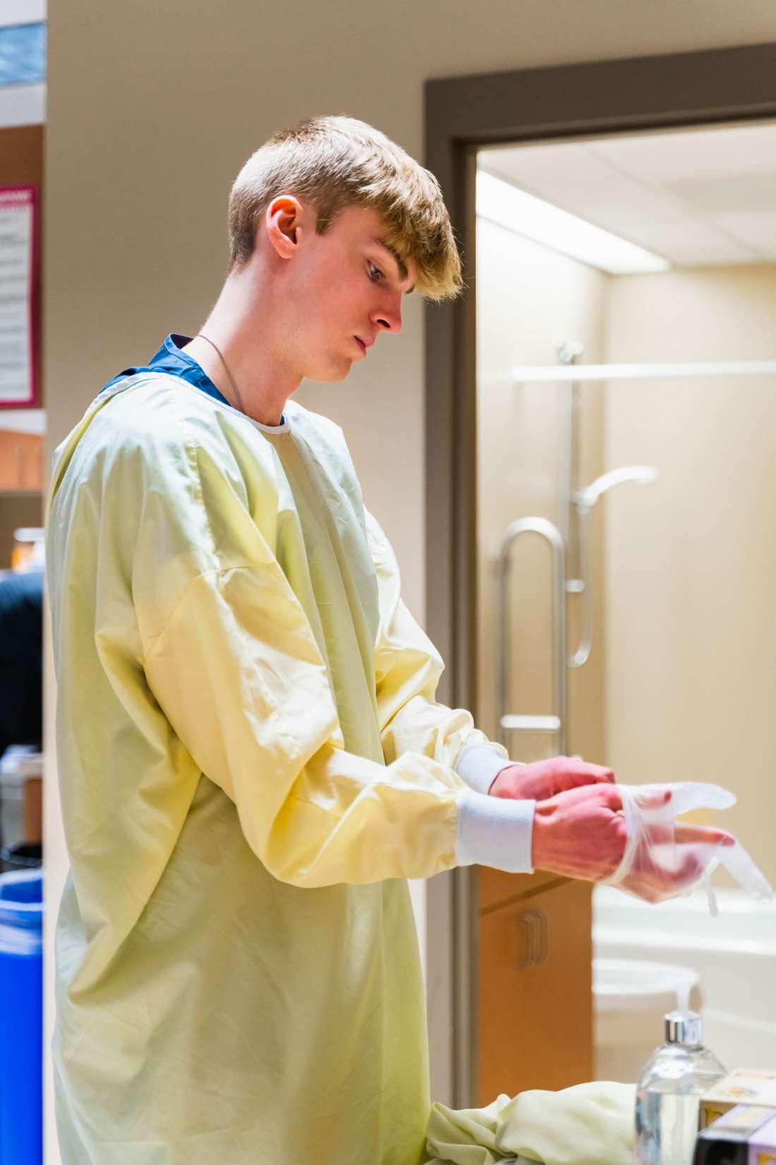 A Nursing Assistant wearing yellow scrubs is putting on rubber gloves.