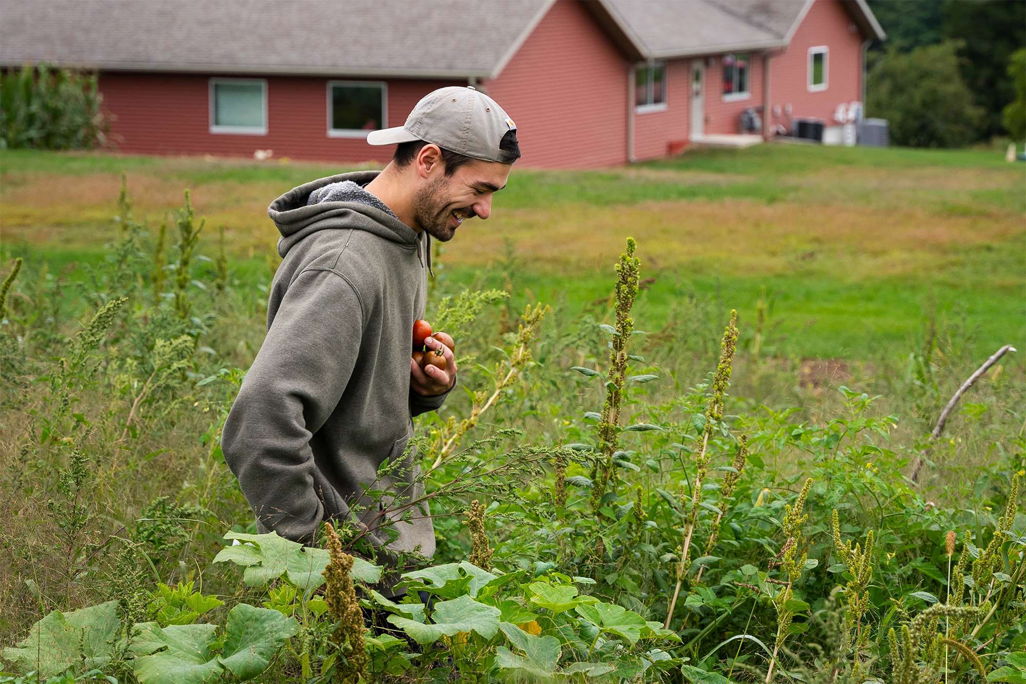 A young man walks through tall grass and weeds holding tomatoes in his hand.