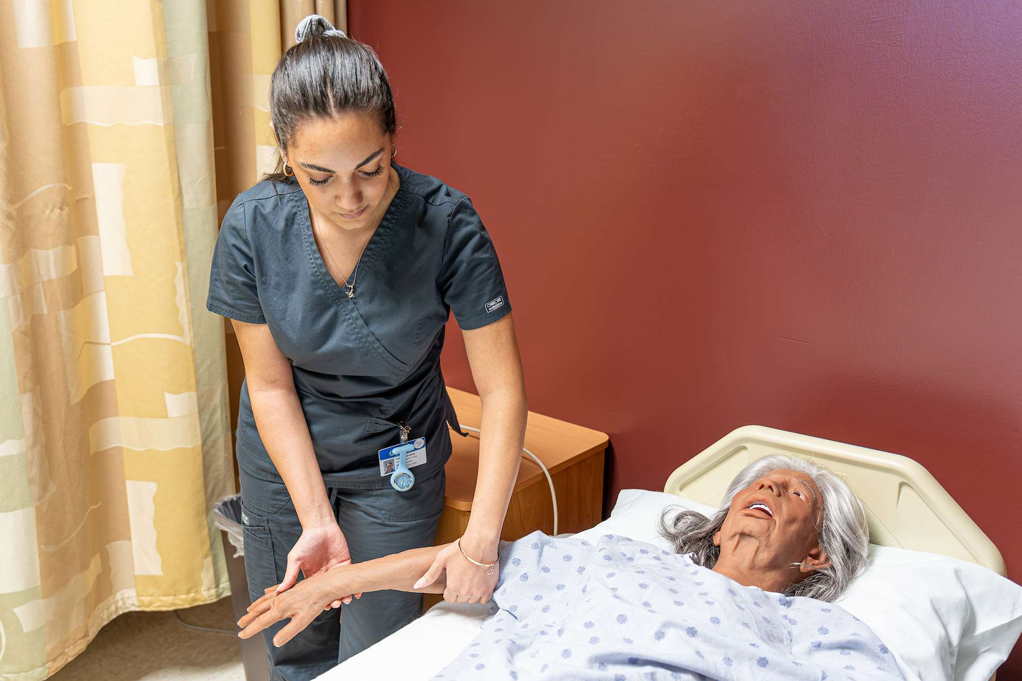 A Certified Nursing Assistant stands next to a hospital bed while moving the right arm of an elderly mannequin.