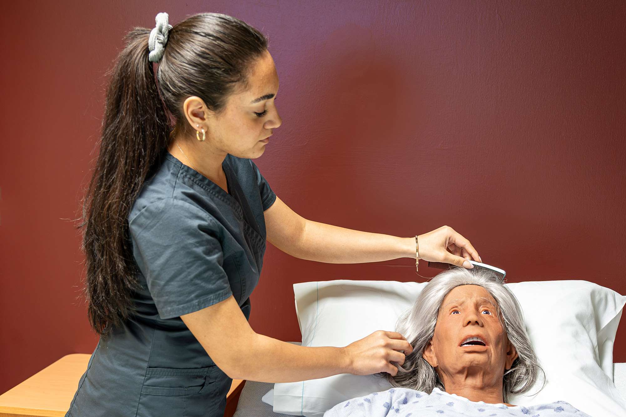 A Certified Nursing Assistant uses a silver colored comb to brush the hair of an elderly mannequin.