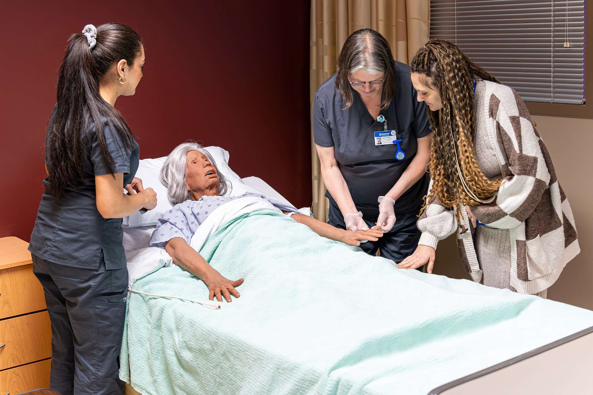 Two Certified Nursing Assistants watch how a different CNA files nails using an elderly mannequin.