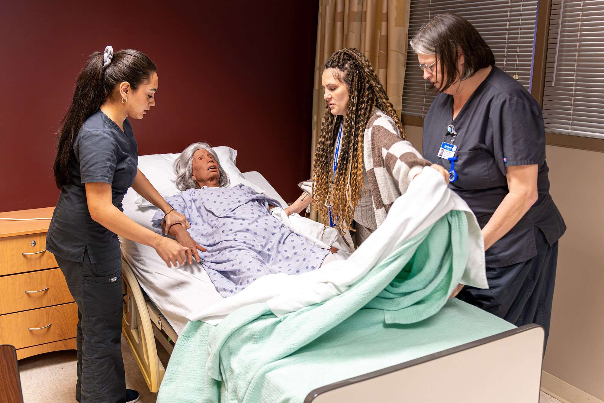 Three certified nursing assistants practice taking vitals using a mannequin and are changing the bedding.