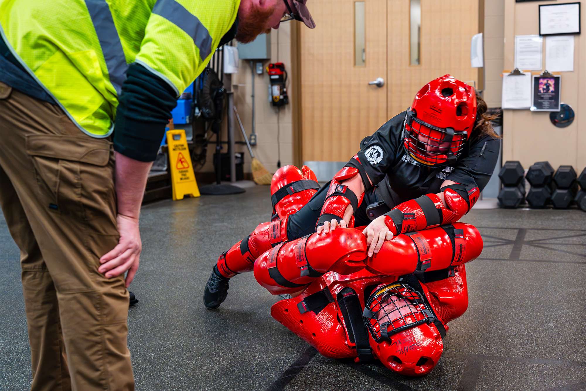 Two people in red protective gear engage in a self-defense training drill, while an instructor observes closely.