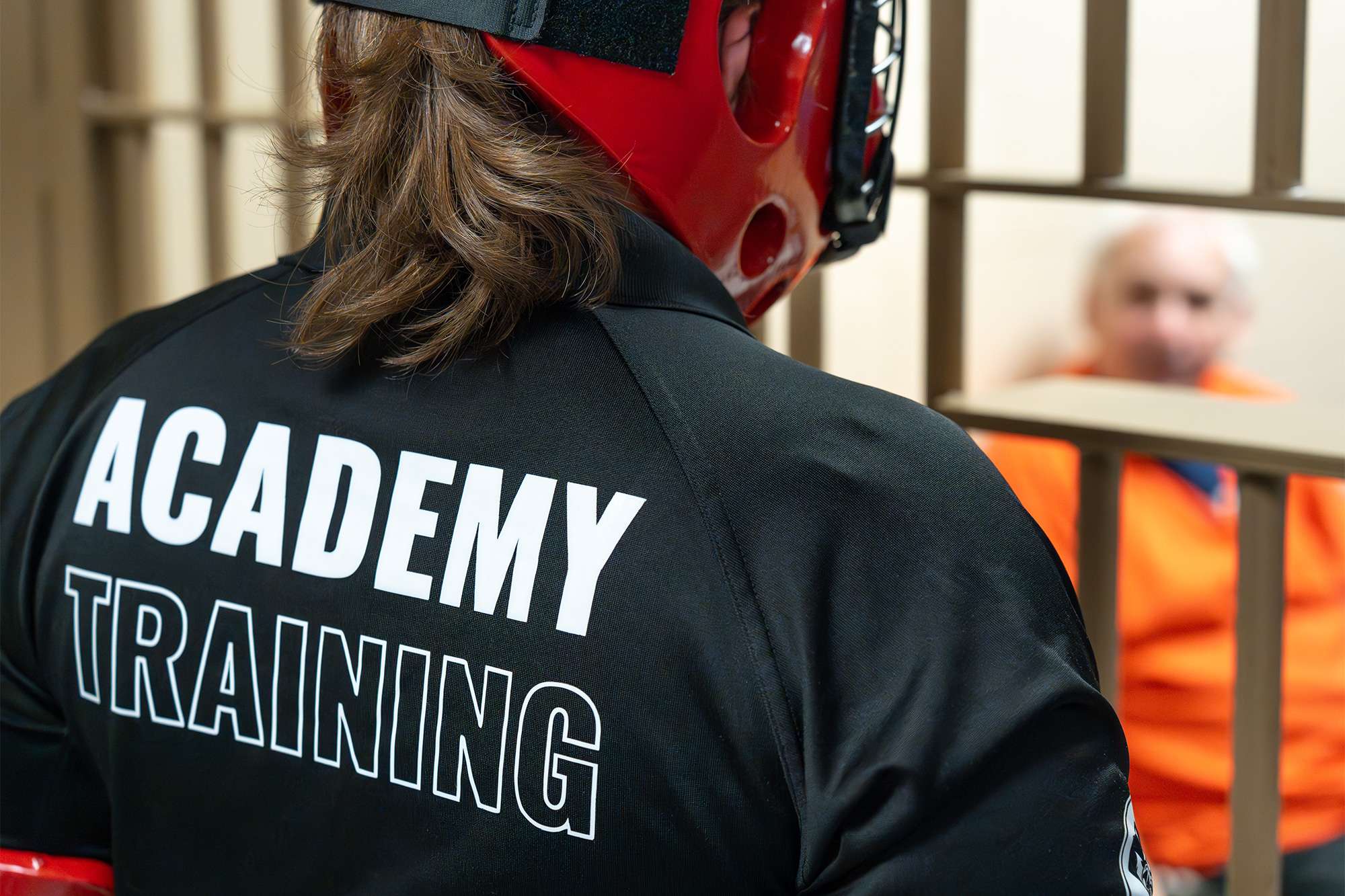A trainee in protective gear with "ACADEMY TRAINING" on their jacket faces an inmate in an orange jumpsuit behind jail bars.