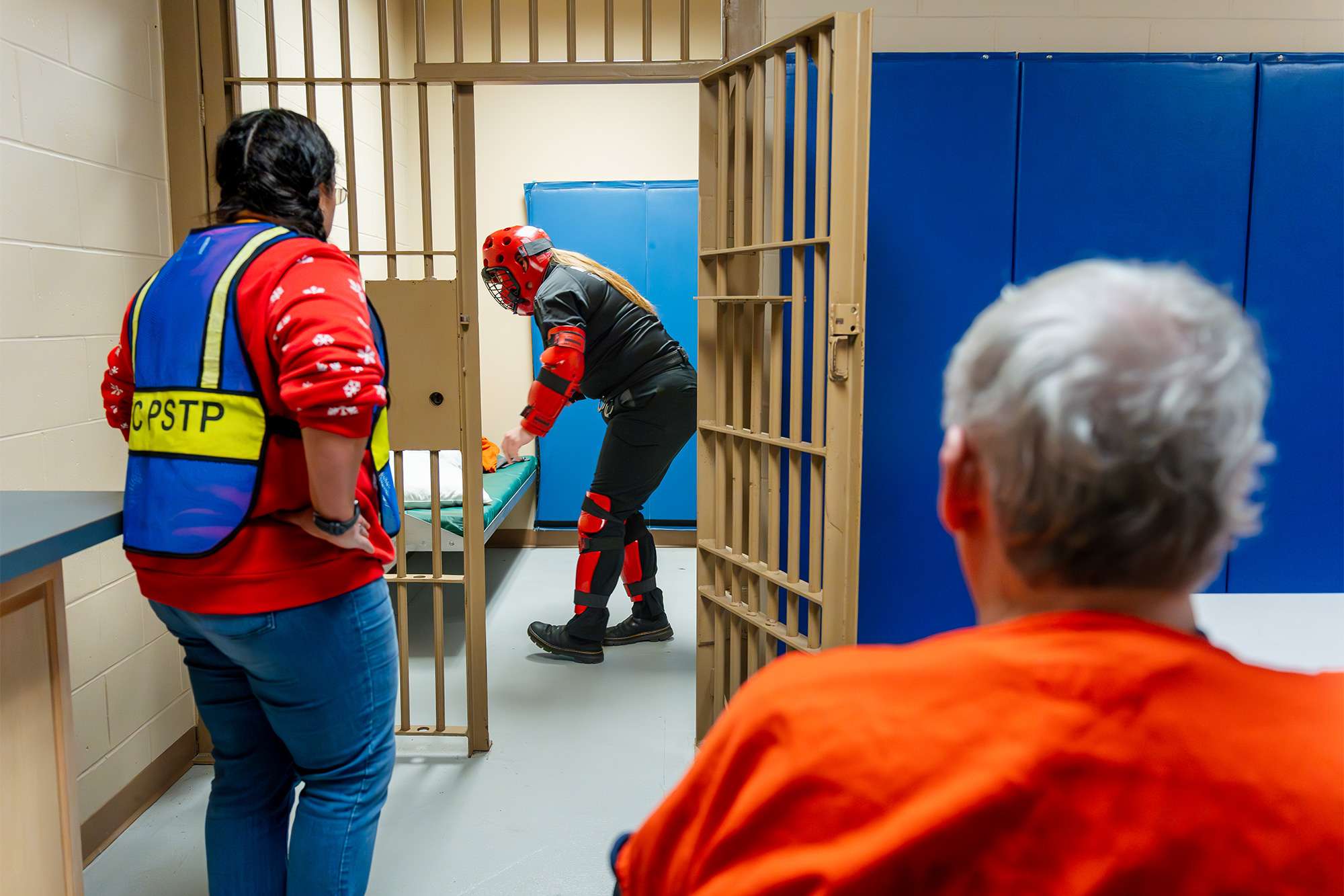 A trainee in red protective gear searches a jail cell while an instructor and an inmate observe the exercise.