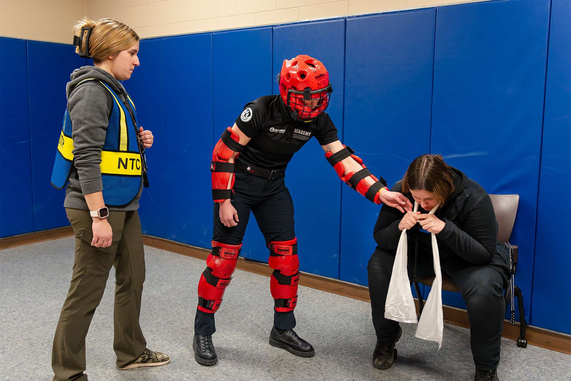 A trainee in red protective gear intervenes as a seated person simulates distress, while an instructor observes the scenario.