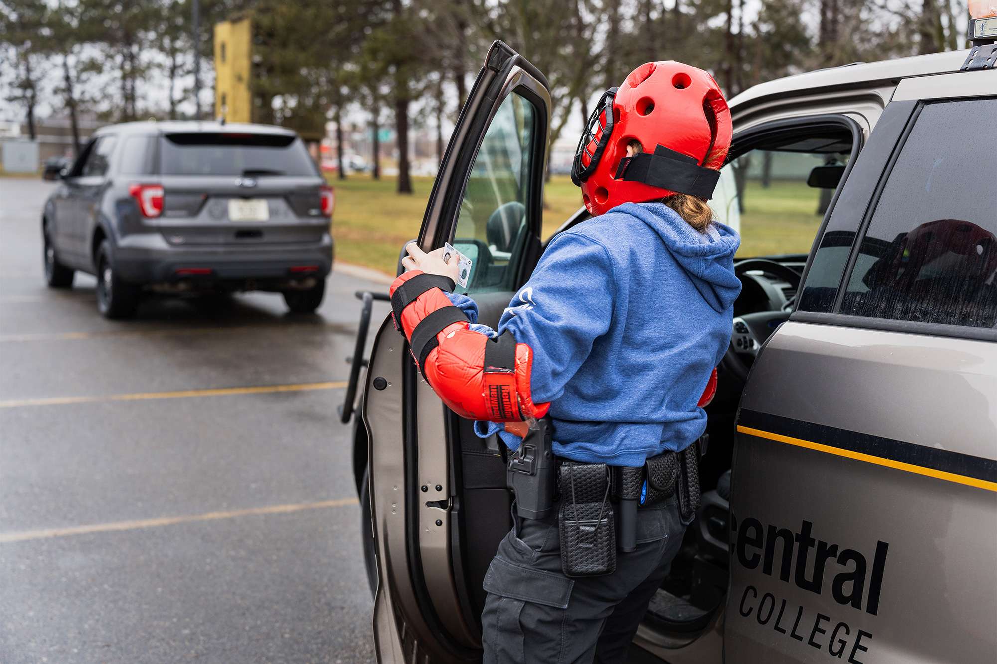 A trainee in red protective gear exits a police vehicle while holding an ID, during a law enforcement training exercise.