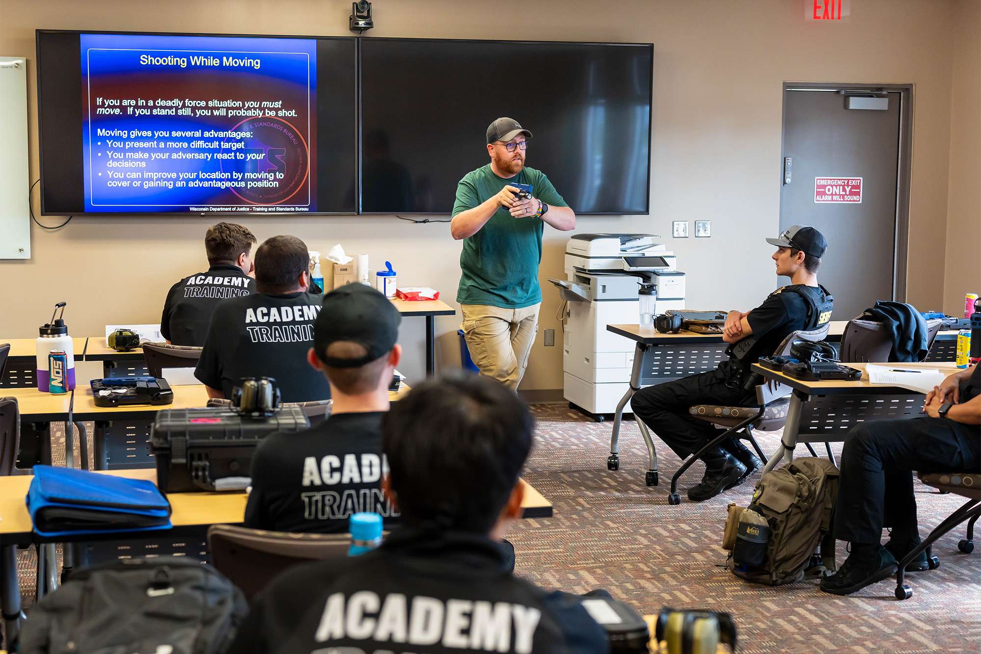 An instructor demonstrates firearm handling to police academy trainees during a classroom session on shooting while moving.