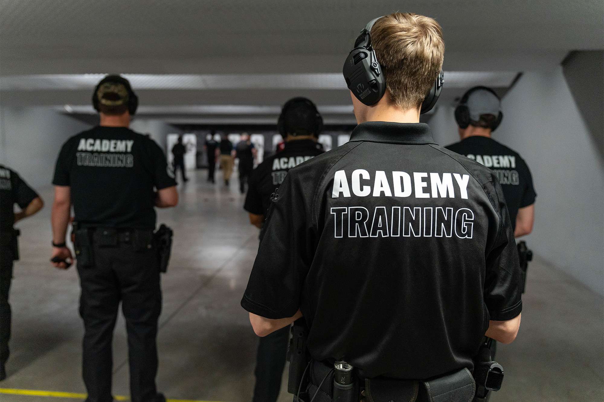Police academy trainees in uniform stand at a shooting range, observing targets during a firearms training session.