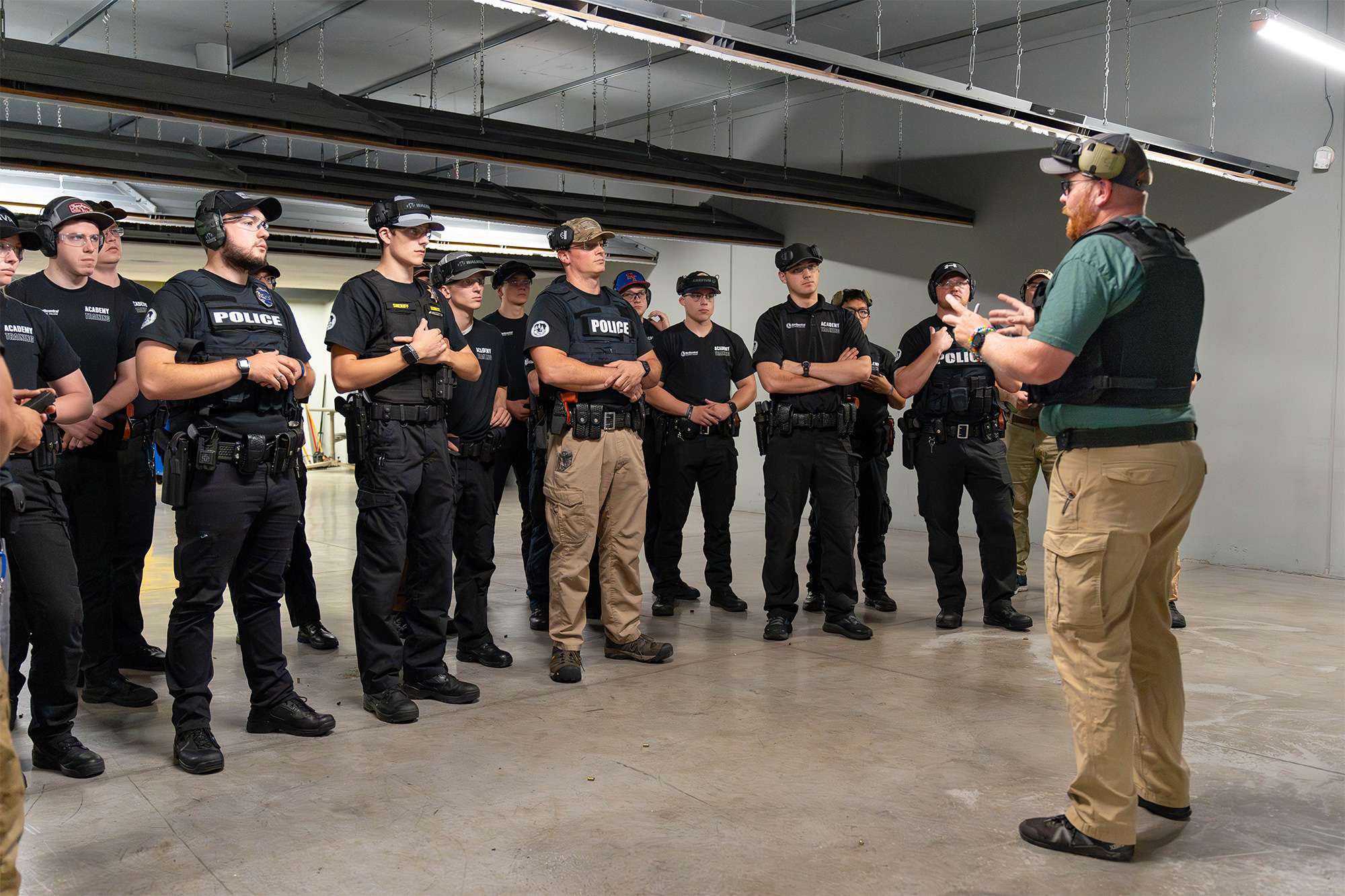 A group of police academy trainees in tactical gear attentively listen to an instructor during a training session indoors.