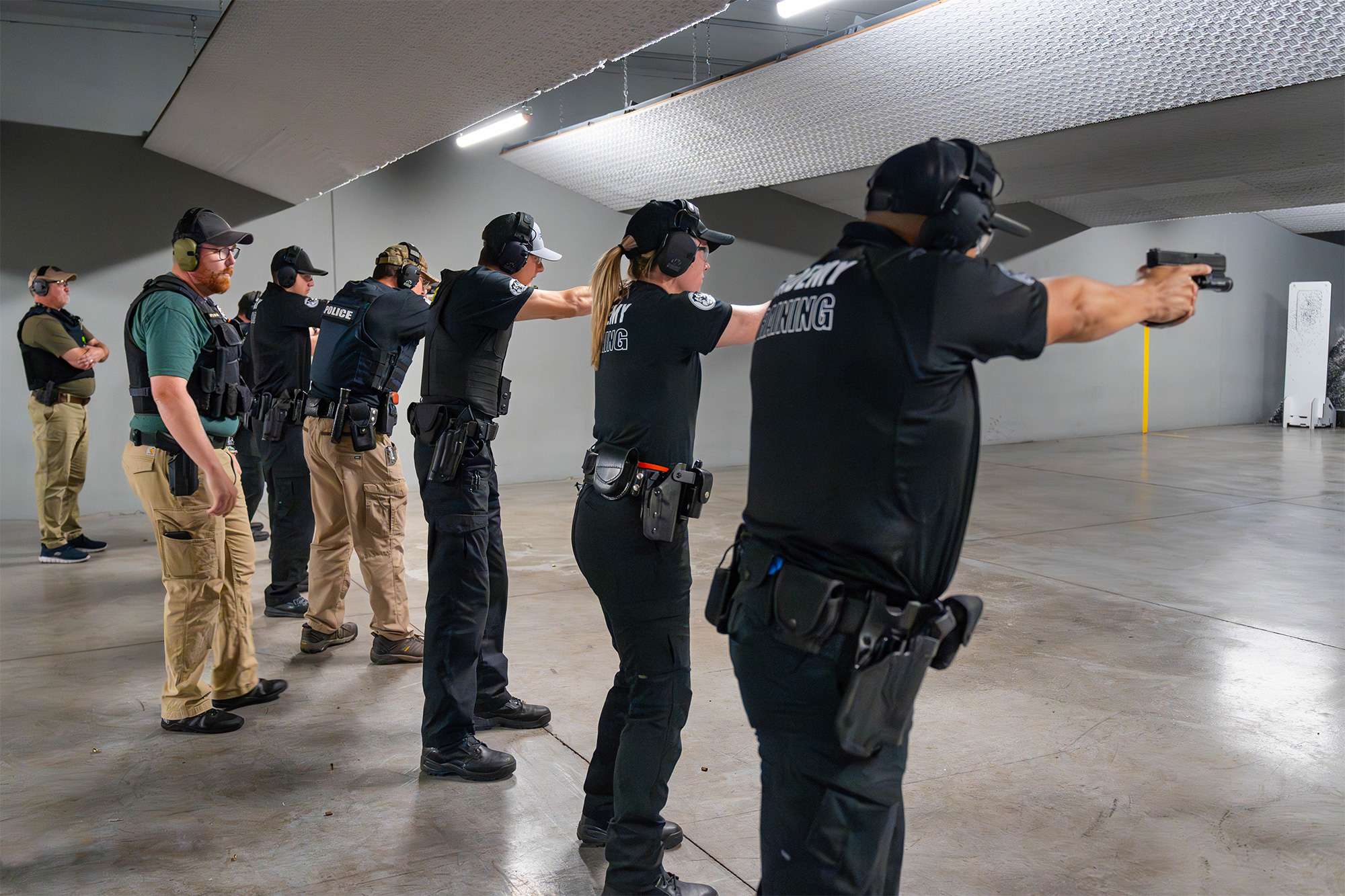 Police academy trainees in uniform practice shooting at a firing range under the supervision of instructors.