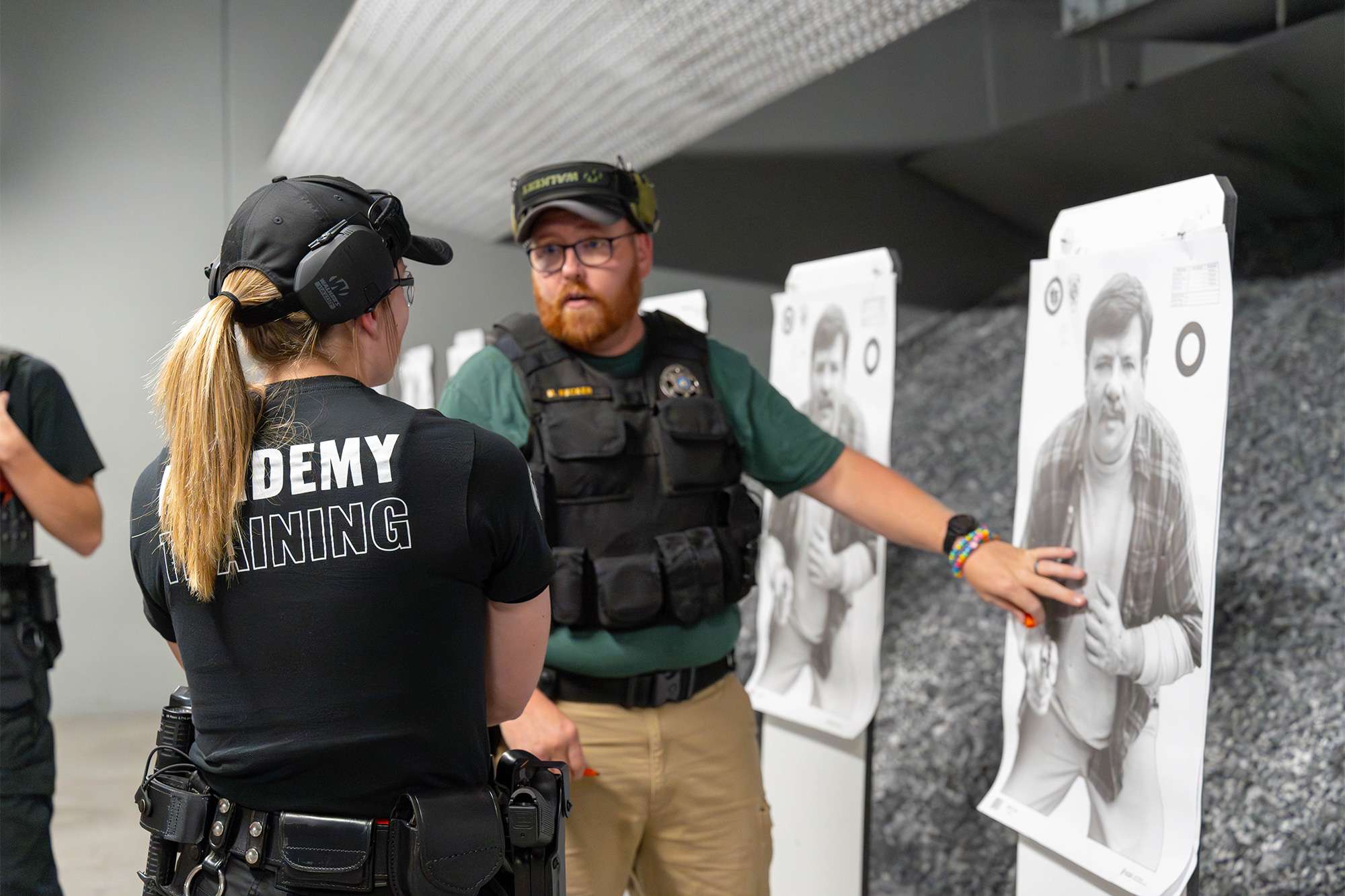 An instructor in tactical gear gives feedback to a police academy trainee at a shooting range, pointing at a target sheet.
