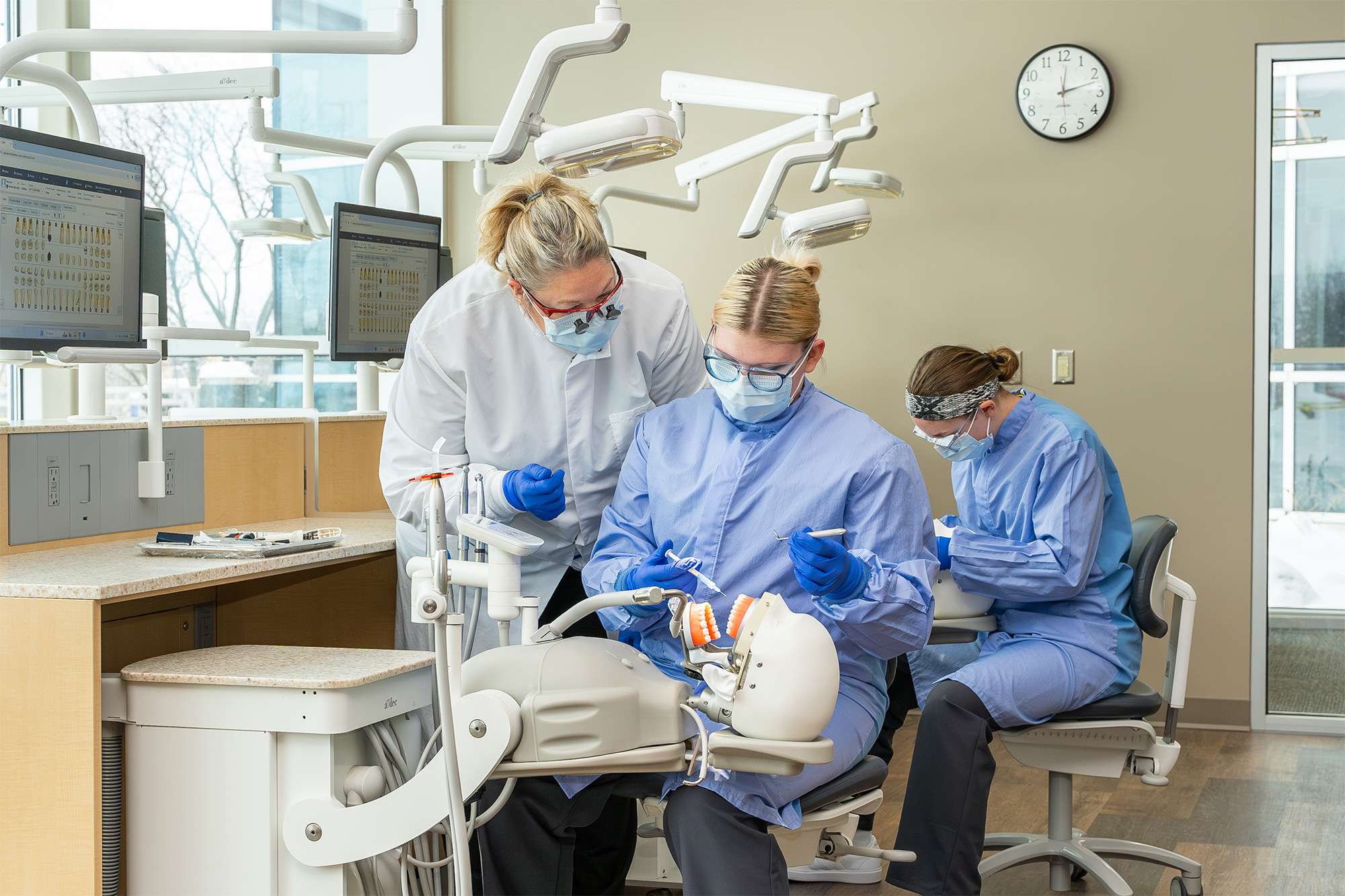 Dental instructor in a white coat guiding a student in blue scrubs practicing on a manikin, with another student nearby.