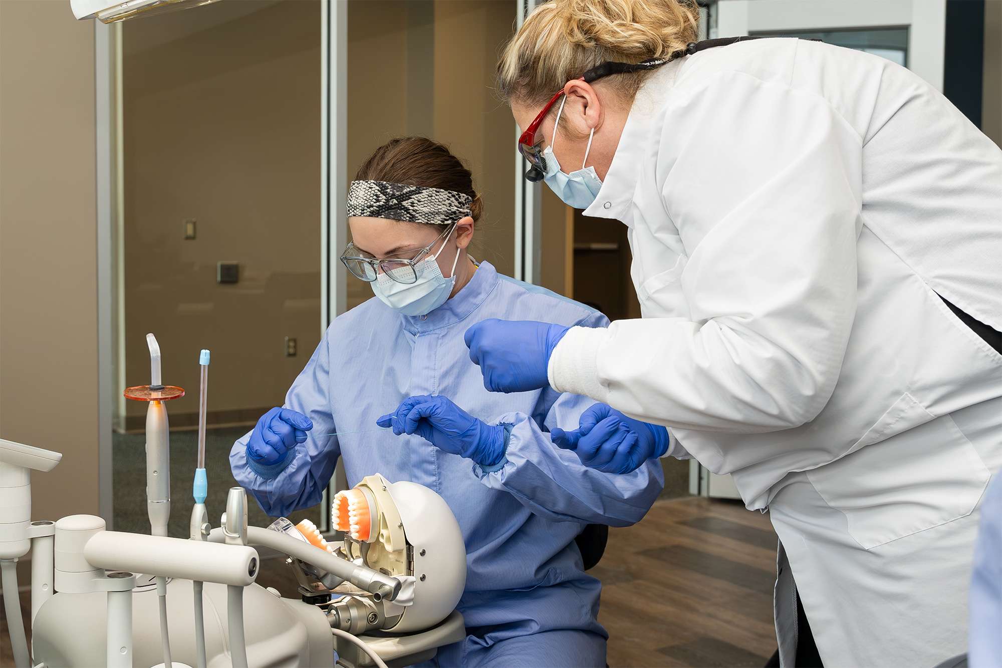 Dental instructor in white coat guiding a student in blue scrubs with hands-on training using a dental mannequin.