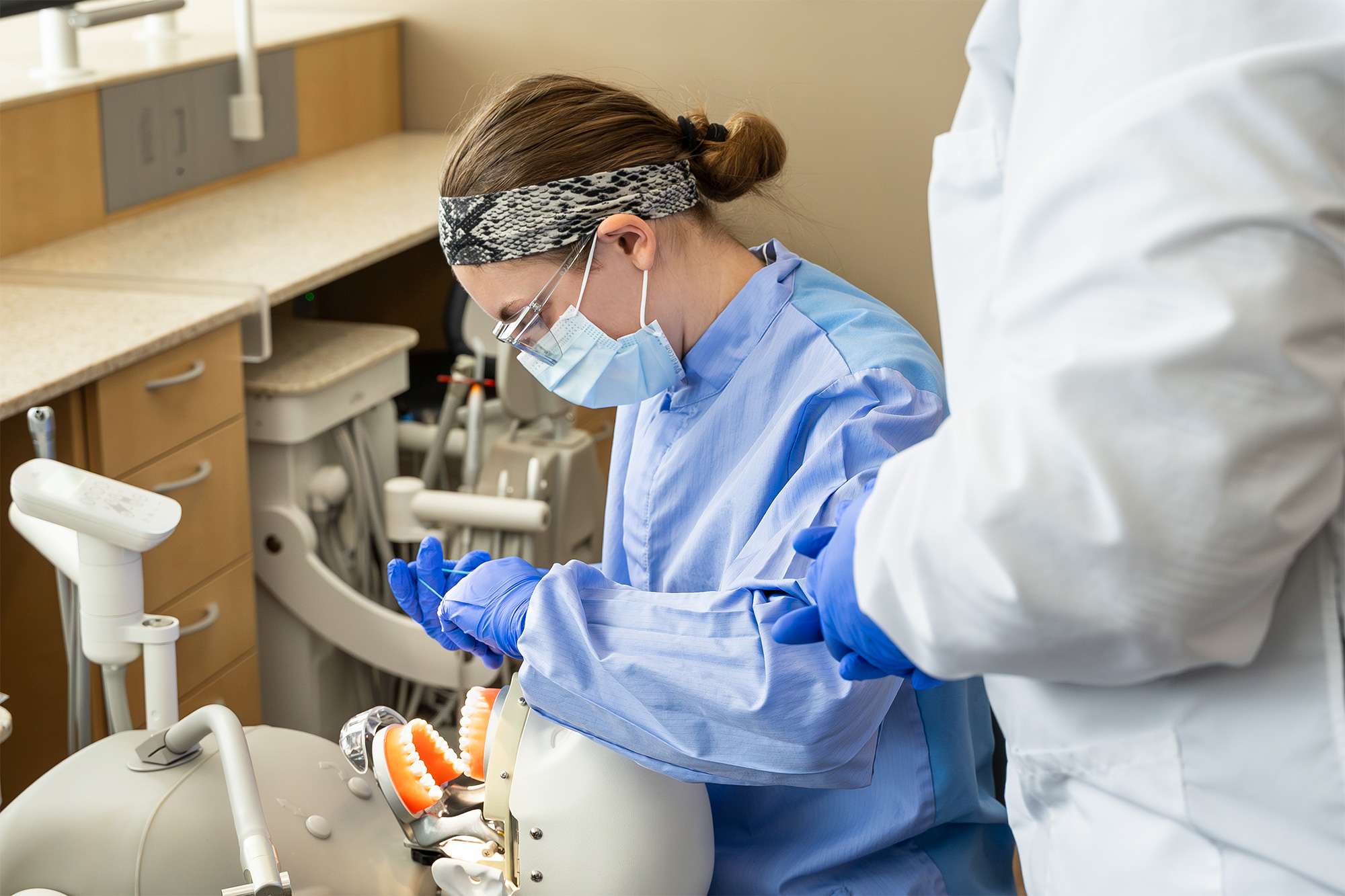 Dental student in blue scrubs practicing on a mannequin, wearing mask and gloves in a clinical setting.