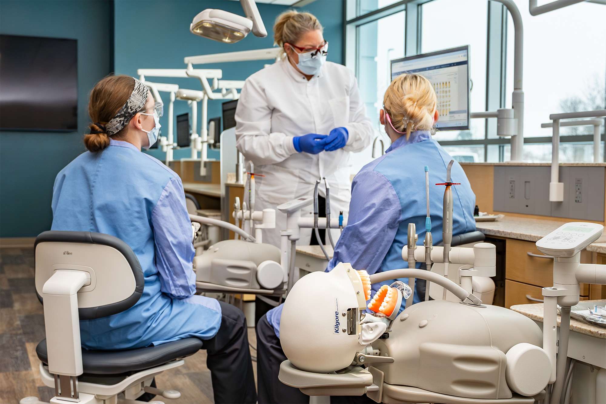 Dental instructor in white coat demonstrating techniques to two students in blue scrubs in a modern dental clinic.