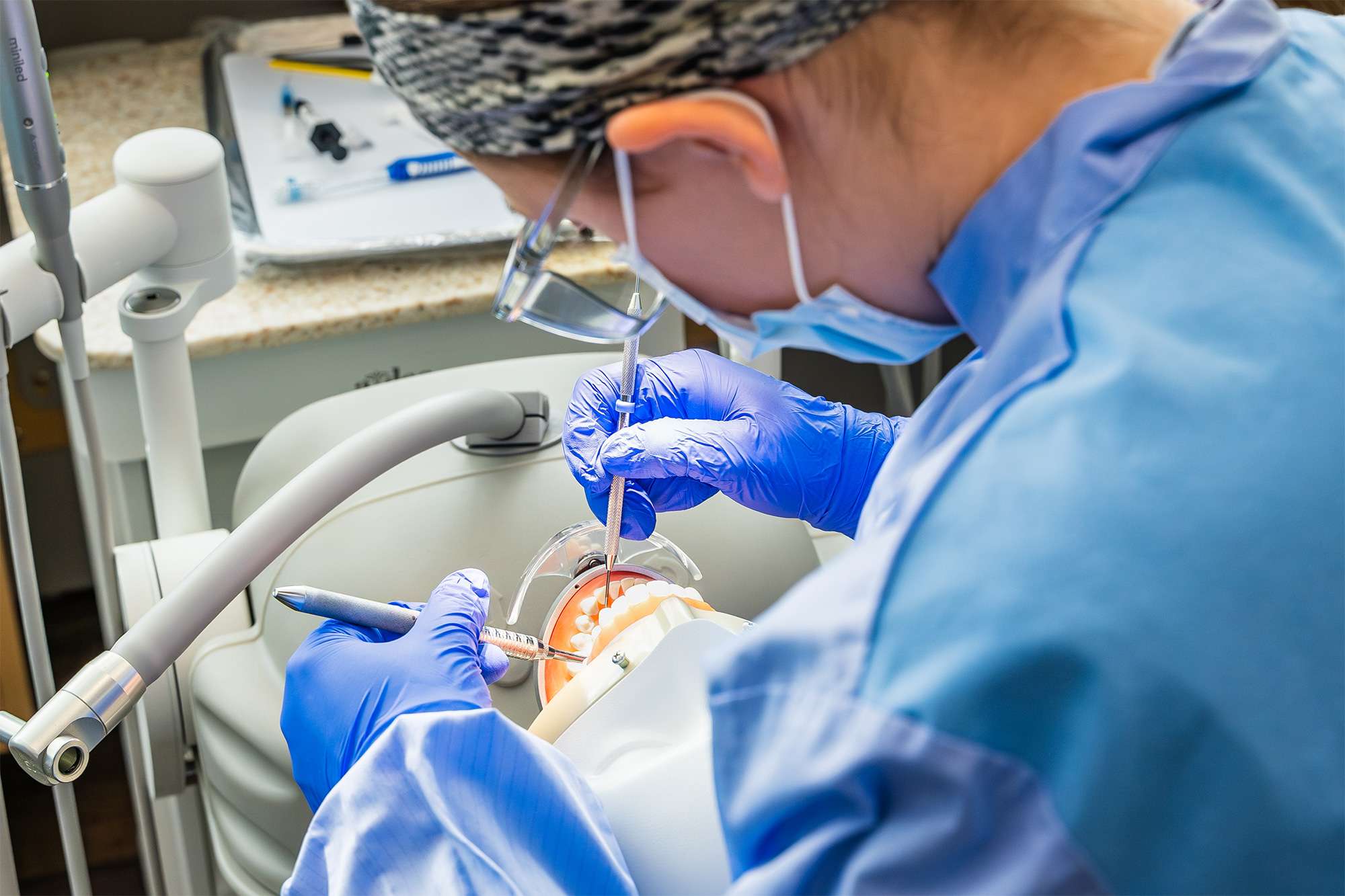 Dental student in blue scrubs and gloves practices procedures on a manikin, using dental instruments in a training lab.