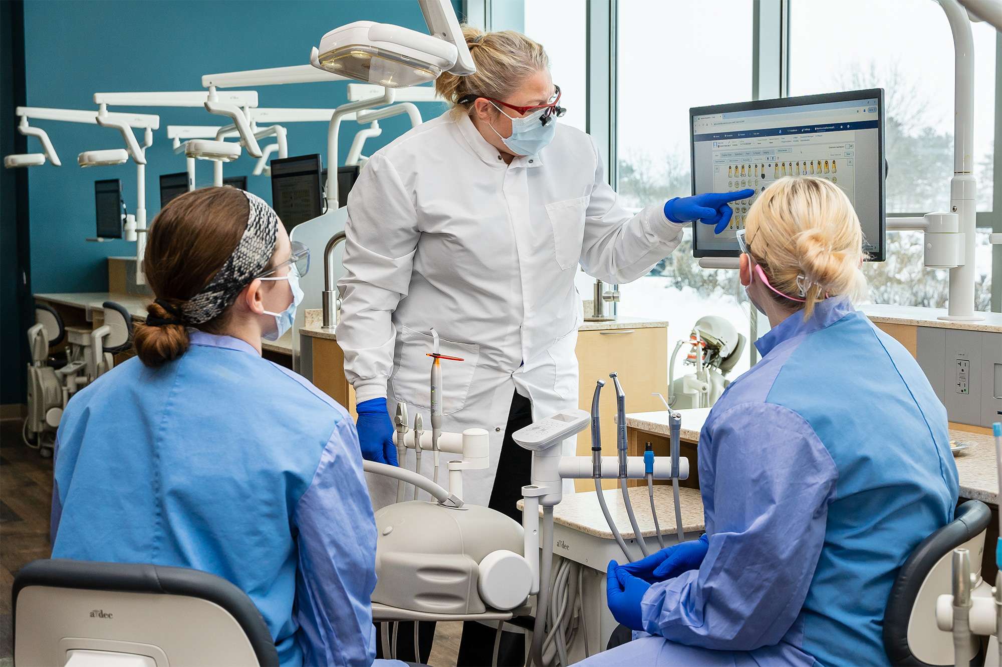 Dental instructor in a lab coat teaching two students in blue scrubs, pointing at a digital dental chart on a screen.