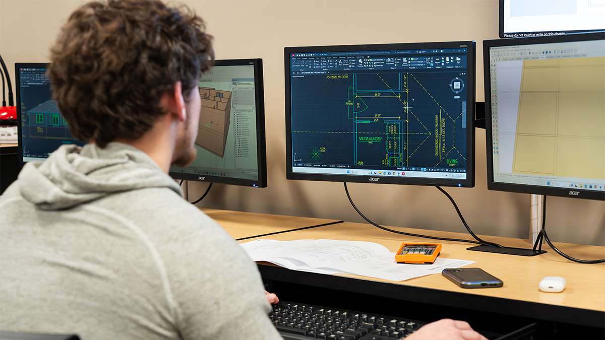 Student works on architectural CAD design using multiple computer monitors in design lab.