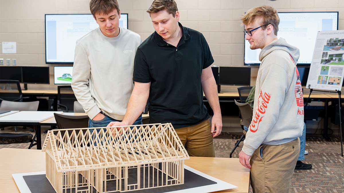 Students examine wooden house frame model in architectural design classroom with computer screens.